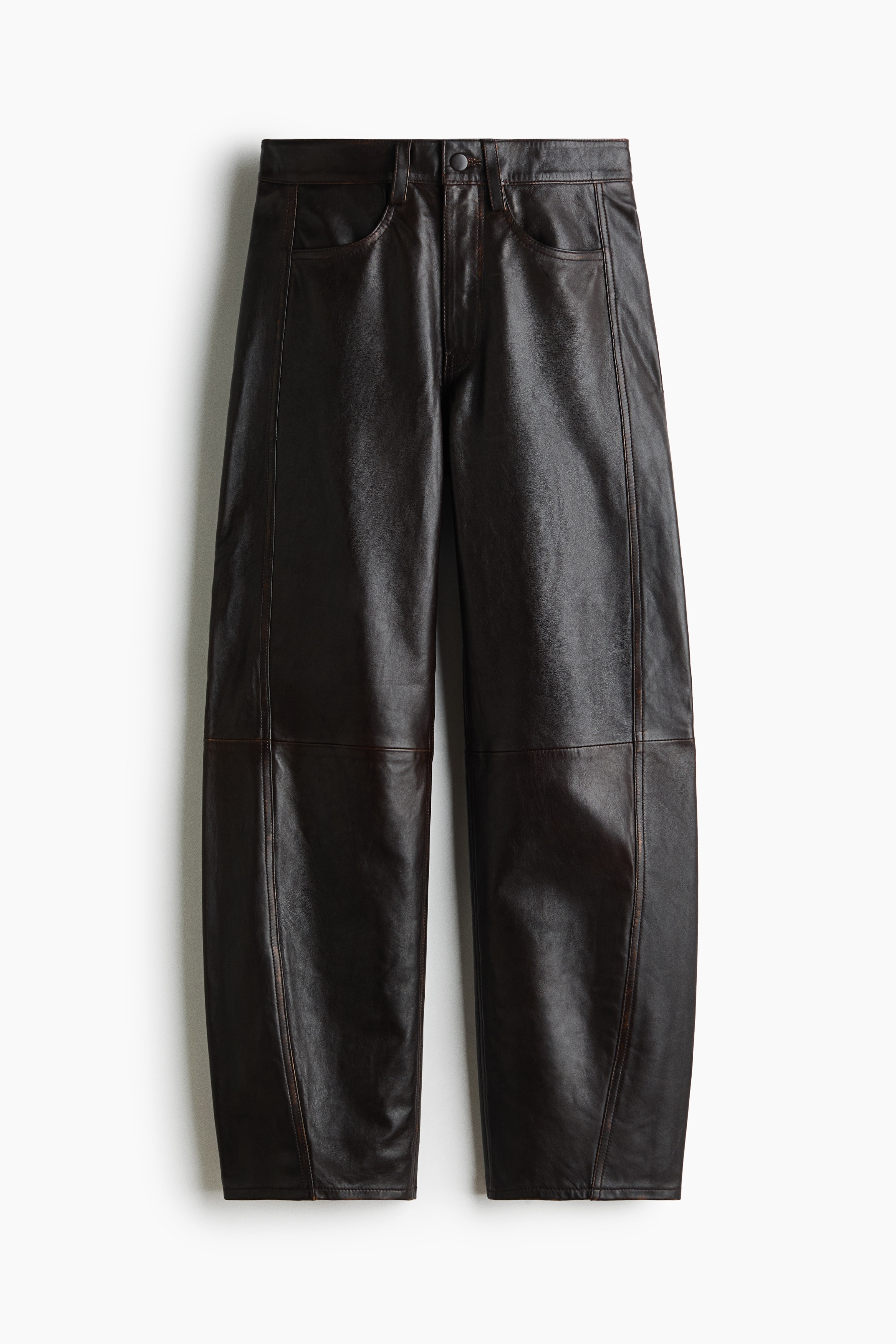 Relaxed-fit leather trousers with a barrel-leg silhouette and prominent shaping seams are being worn by the model, styled with a black leather belt. The model is standing with one hand resting in the front pocket of the trousers, which are paired with a dark floral jacket and black pointed-toe boots.