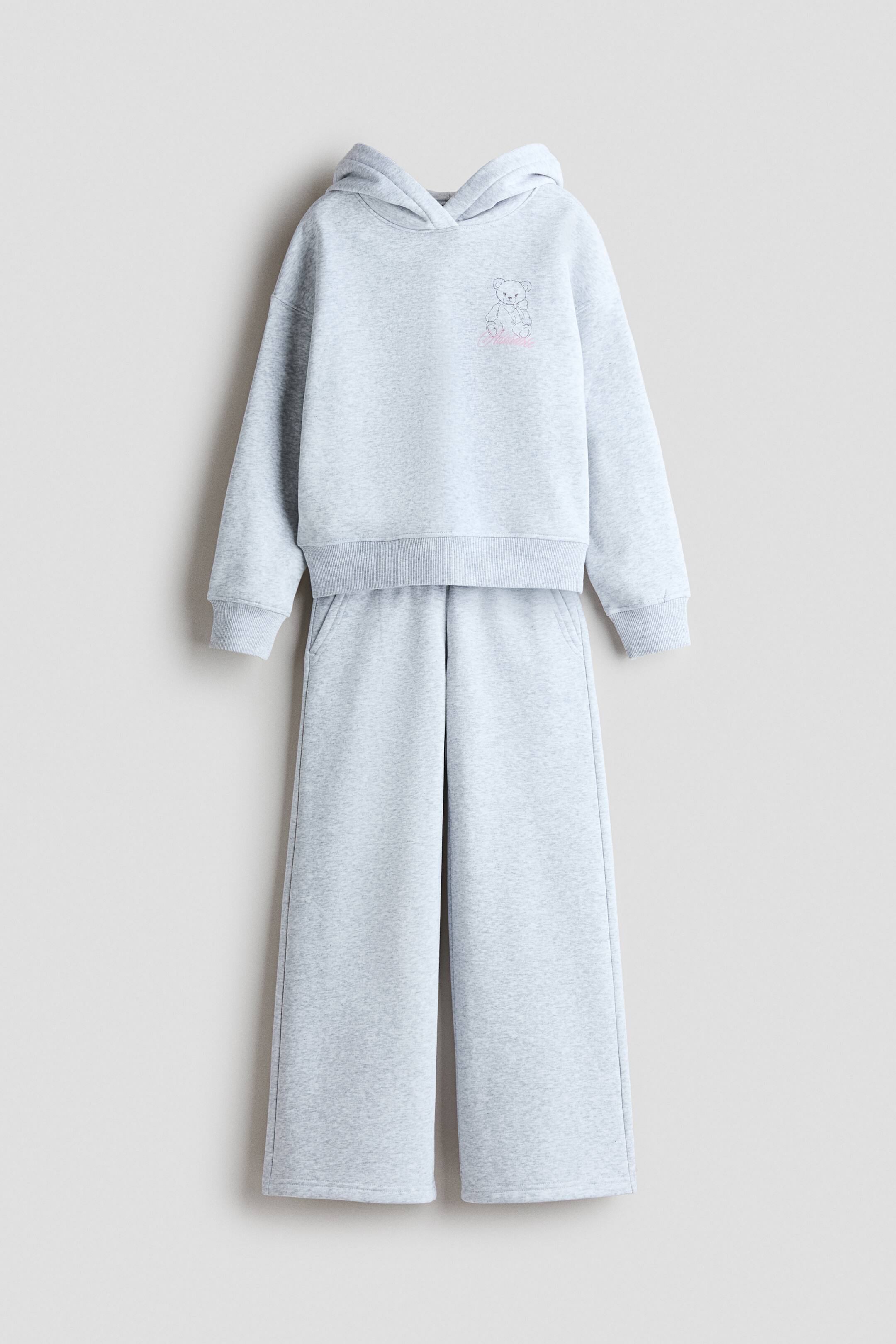 查看大圖: 一套淺灰色兩件式家居服以前視角懸掛展示,包含連帽上衣與同色寬褲運動褲。寬鬆版型的連帽上衣採交叉式帽兜、羅紋袖口,左胸處有一個小熊圖案;運動褲則呈現寬褲剪裁,並設有側口袋。