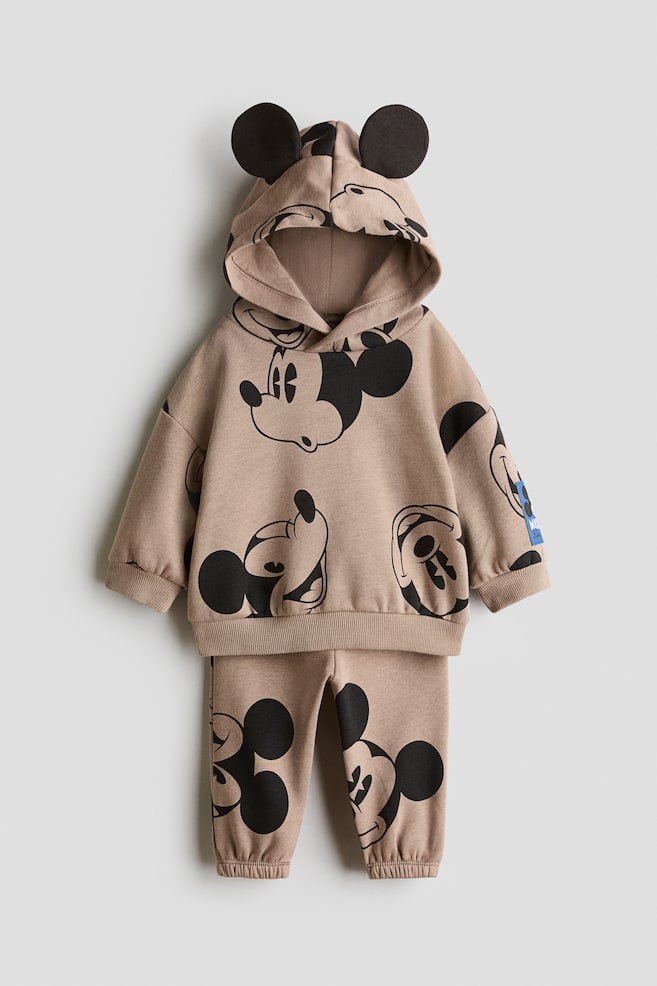 This two-piece hoodie and track pants set is shown from the front, featuring a light brown fabric with an all-over black Mickey Mouse print. The hoodie features an attached hood with black 3D ears, dropped shoulders, and ribbed trims, while the matching track pants feature elasticated ankle cuffs. - 1