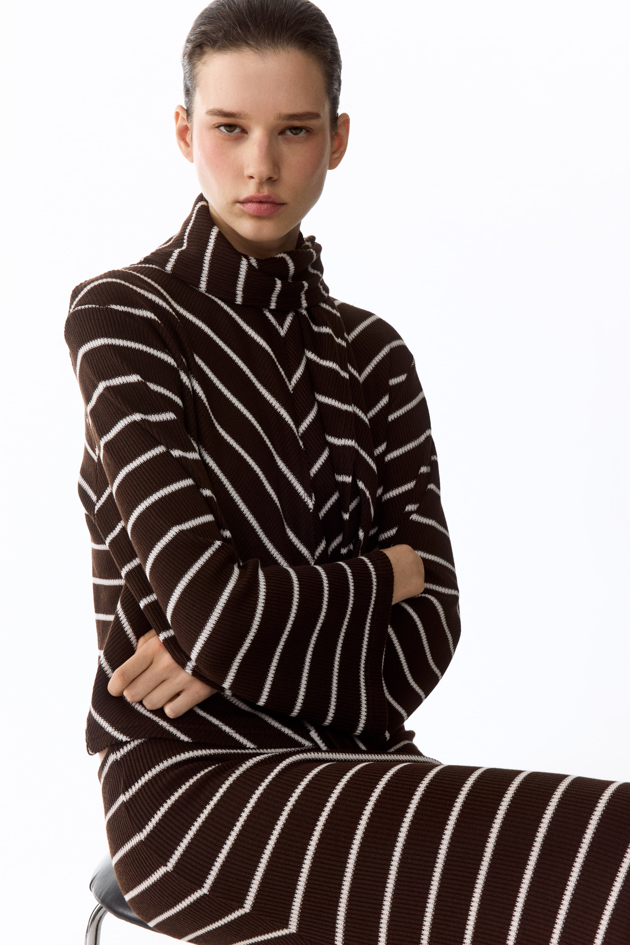 Ver imagem maior: Um conjunto de malha canelada às riscas castanhas e brancas, composto por uma camisola de gola alta e uma parte inferior coordenada. A camisola apresenta riscas diagonais brancas formando um padrão em chevron no peito e nas mangas, enquanto a parte inferior exibe riscas diagonais consistentes.