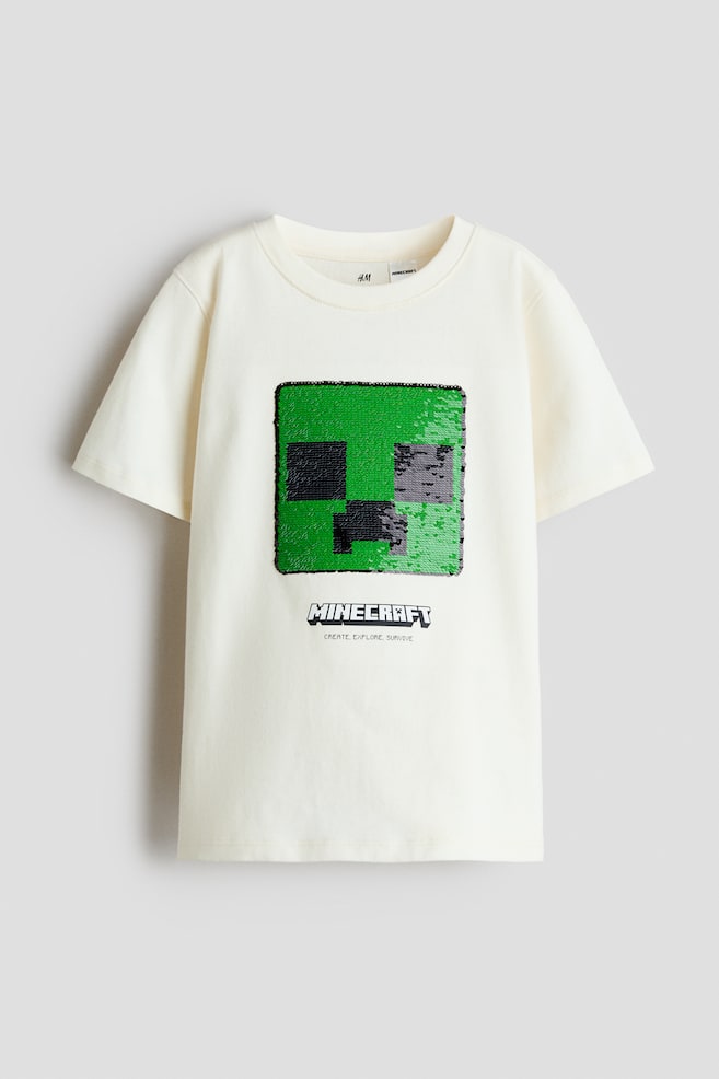 A front-facing off-white cotton jersey t-shirt is featuring a ribbed crew neckline, short sleeves, and a straight hem. The center front is displaying a square, green and black sequined Minecraft Creeper face with the "MINECRAFT" logo and slogan printed directly below it. - 1