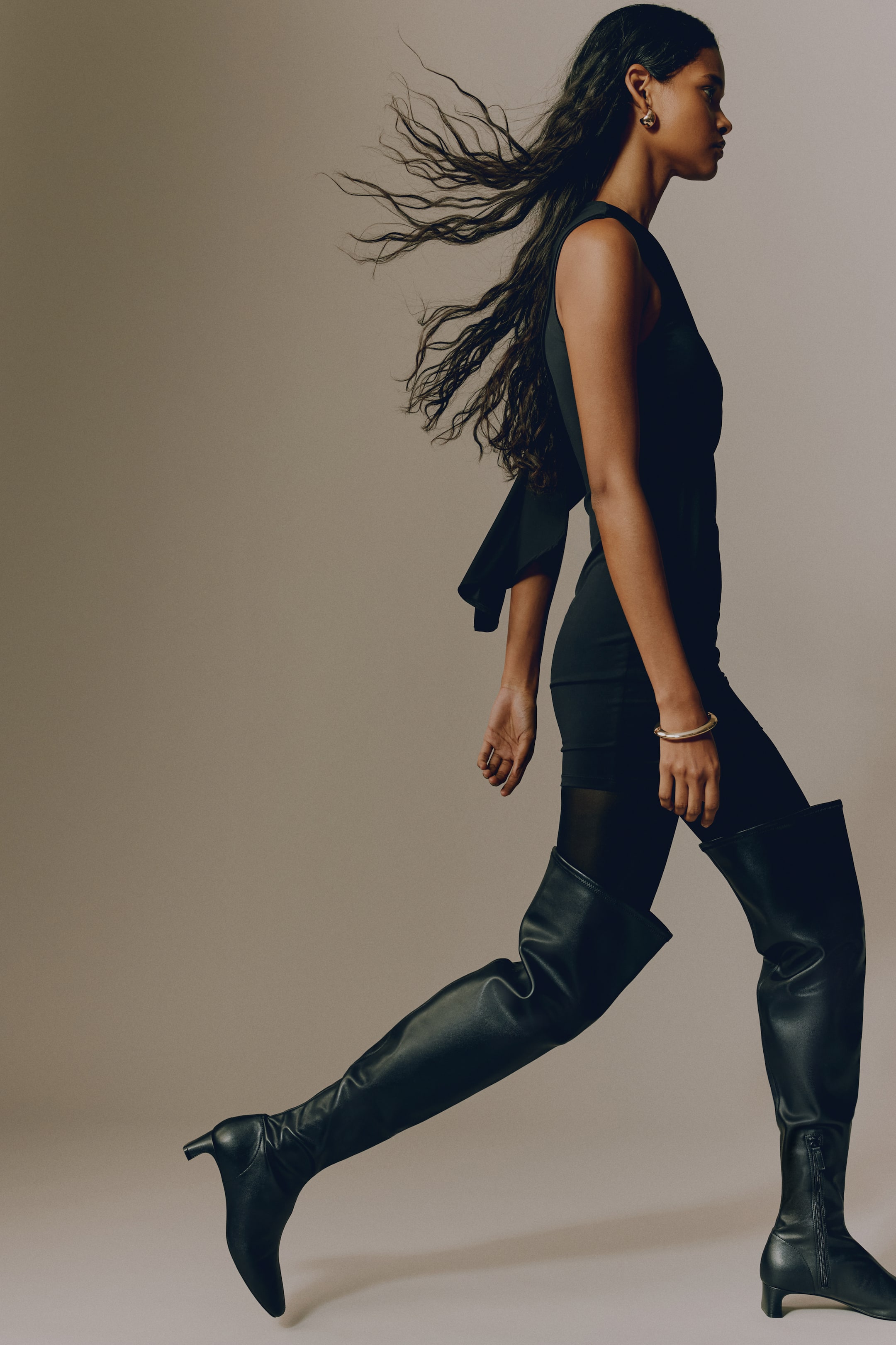 View larger image: The model is wearing a black mini dress with a cape detail. A flowing cape extends from one shoulder of the dress as the model walks, and the look is styled with black sheer tights and over-the-knee boots.