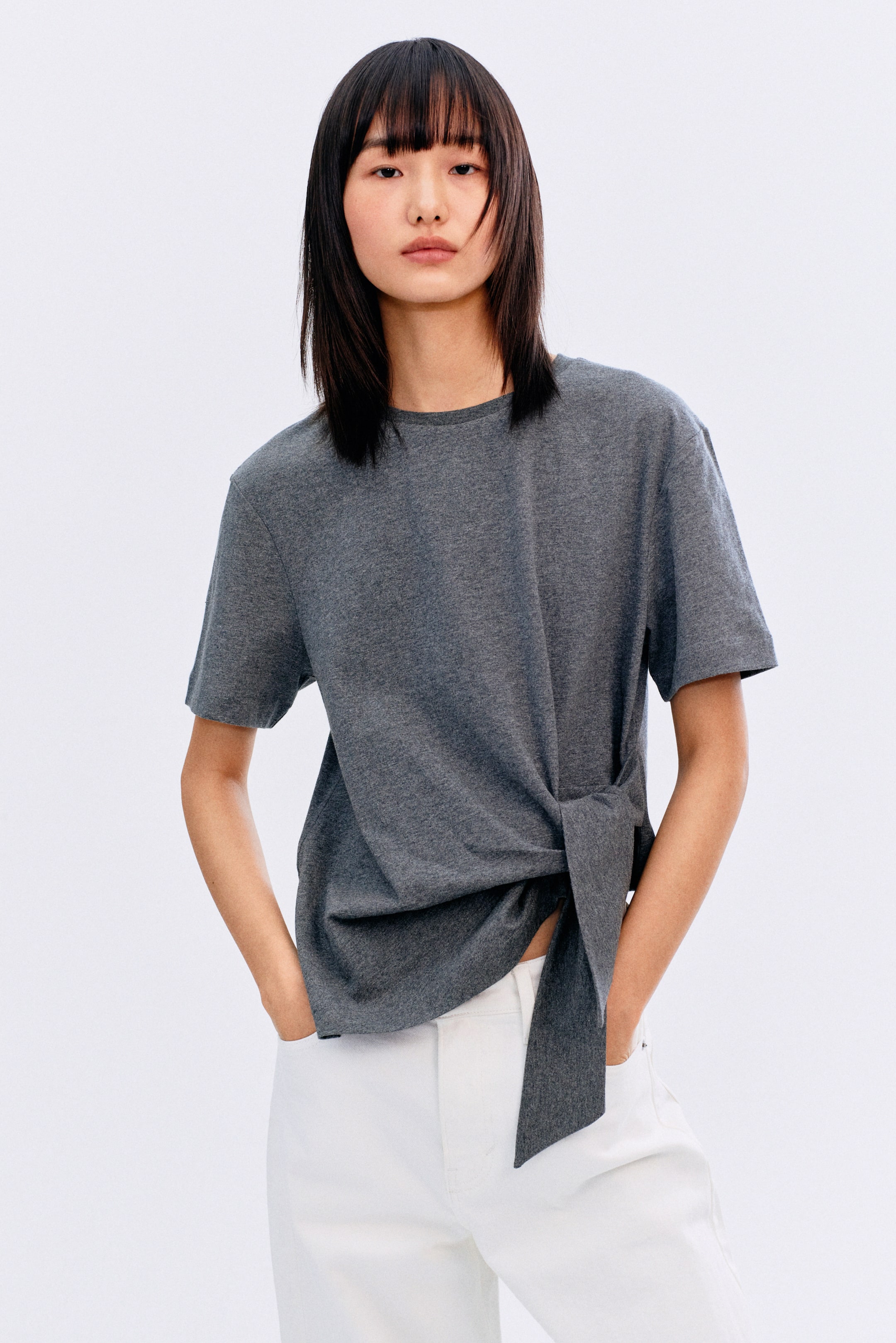 拡大画像を表示: ダークグレーの半袖Tシャツは、クルーネックとリラックスフィットを特徴としており、裾が左側で結ばれています。