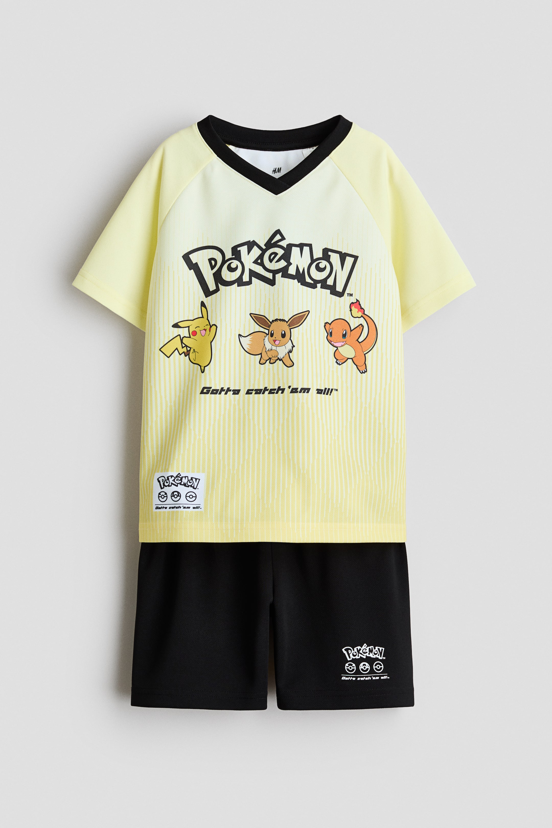 Ver imagen más grande: Un conjunto de camiseta de fútbol amarilla clara y pantalones cortos de fútbol negros muestra su parte delantera. La camiseta presenta un sutil patrón de rayas verticales, un cuello en V con ribetes negros, mangas raglán amarillas claras y un destacado logo de 'Pokémon' con los personajes Pikachu, Eevee y Charmander impresos en el frente, mientras que los pantalones cortos negros cuentan con una cintura elástica y un logo blanco de 'Pokémon' impreso en la pierna izquierda.