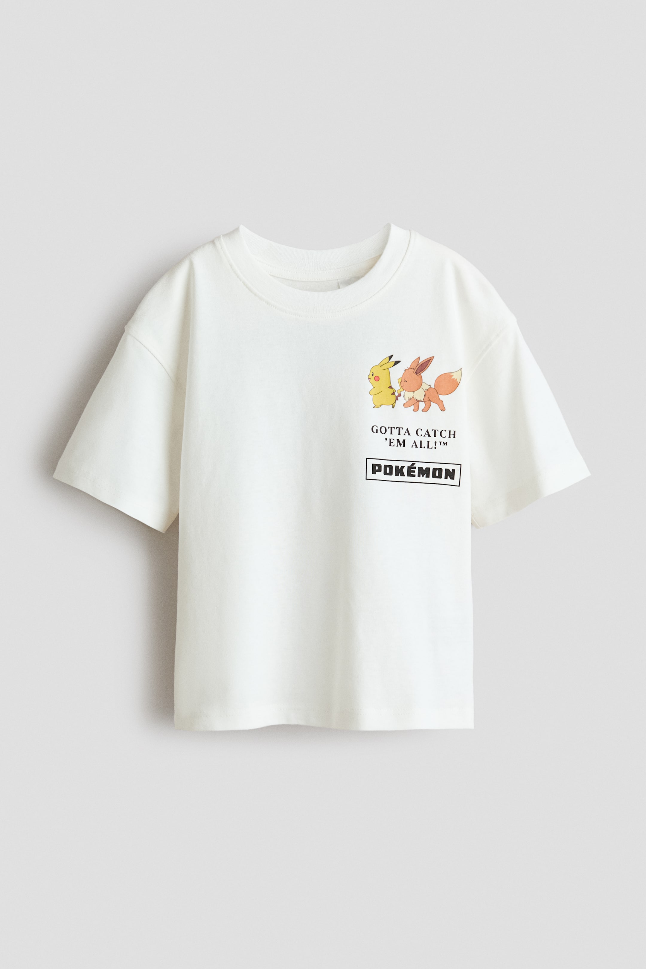 Visa större bild: En off-white, oversized t-shirt vänd framåt, med en ribbad halsringning och nedhasade axlar. Den är tillverkad av mjuk, tryckt bomullsjersey och visar en grafik av Pikachu och Eevee-karaktärer tillsammans med texten 'GOTTA CATCH 'EM ALL!?' och 'P O K É M O N'.