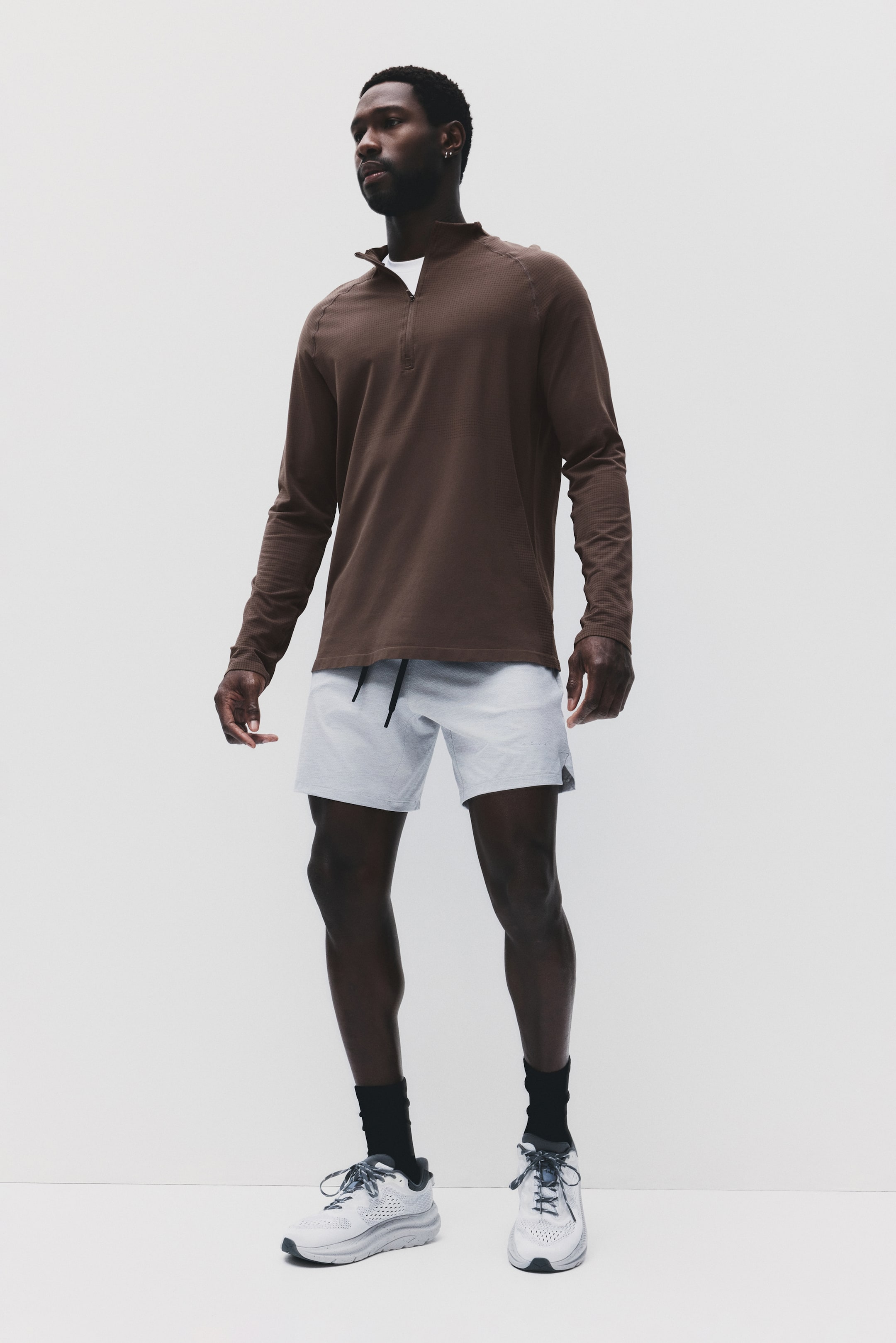 View larger image: The model is wearing a brown muscle fit quarter-zip sports top, showcasing long sleeves and a subtle textured pattern. It's layered over a white t-shirt and paired with light grey shorts.