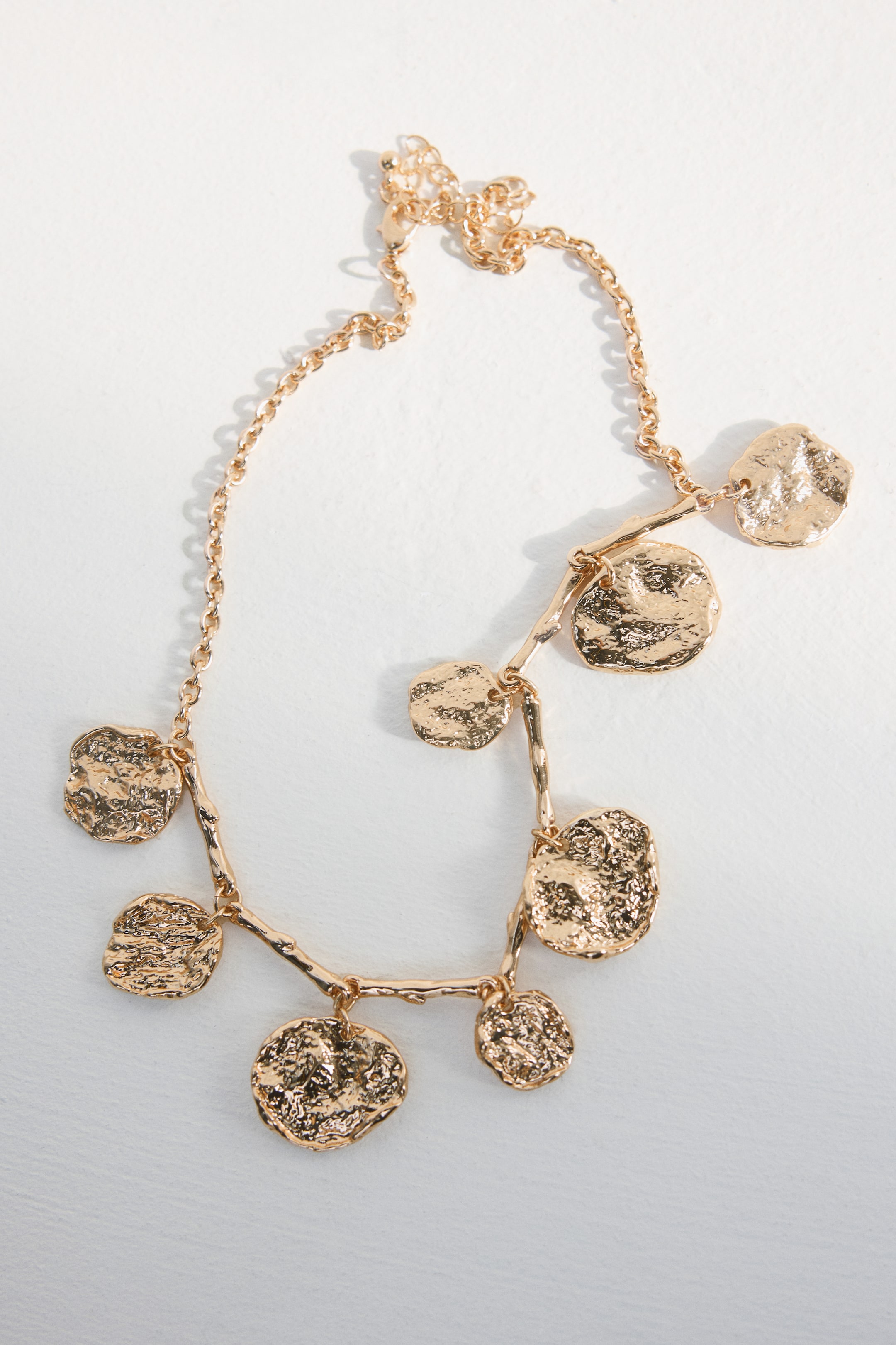 View larger image: A gold-toned necklace is laid flat, showcasing a delicate chain adorned with multiple irregular, hammered disc charms. These charms are linked by organically-shaped connectors, and a lobster clasp with an extender chain is present.