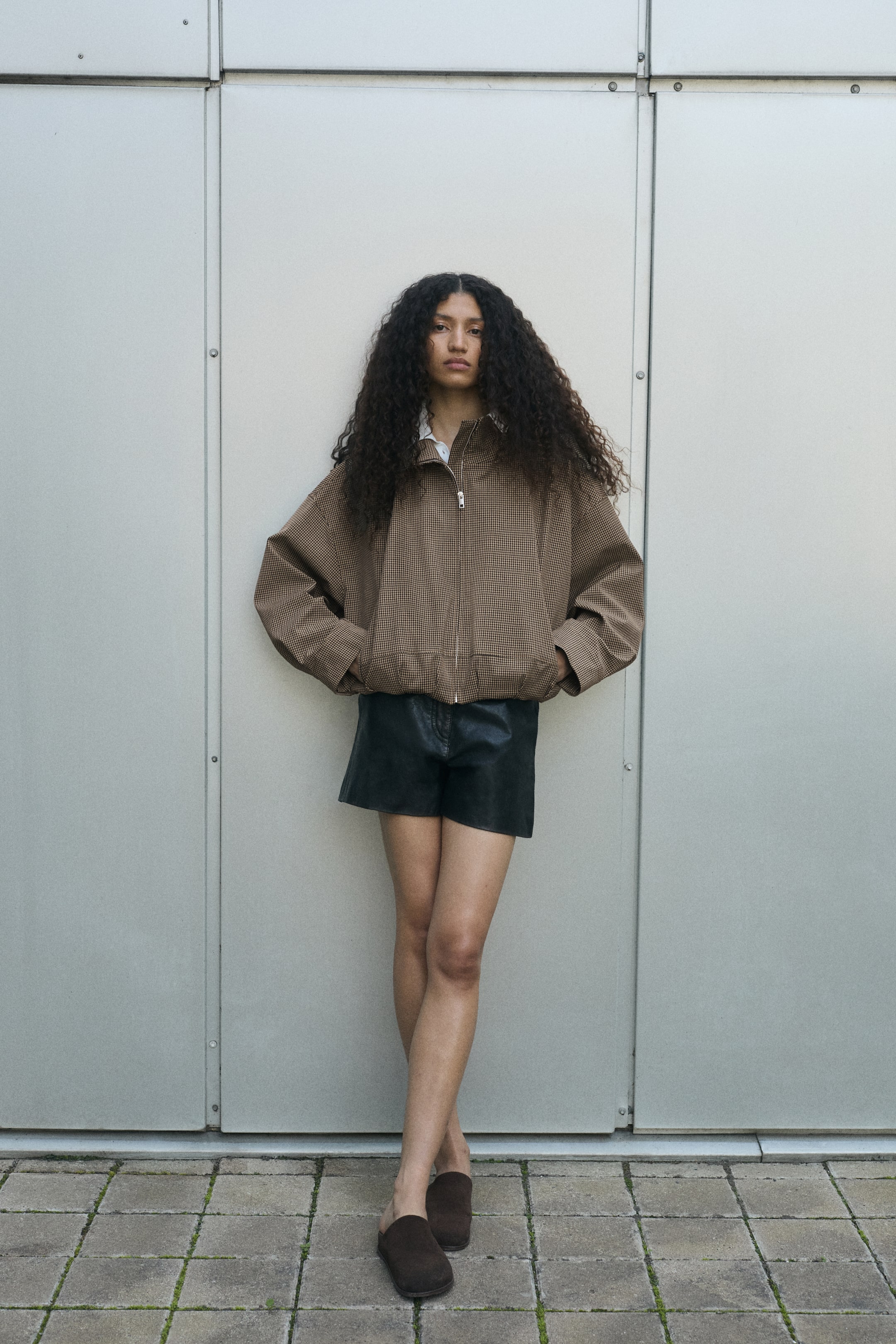 View larger image: The model is wearing a brown patterned oversized jacket with a funnel neck, dropped shoulders, and a gathered bubble hem, with their hands tucked into the jetted pockets. The jacket is zipped up in the front, layered over a white collared shirt, and styled with black shorts.
