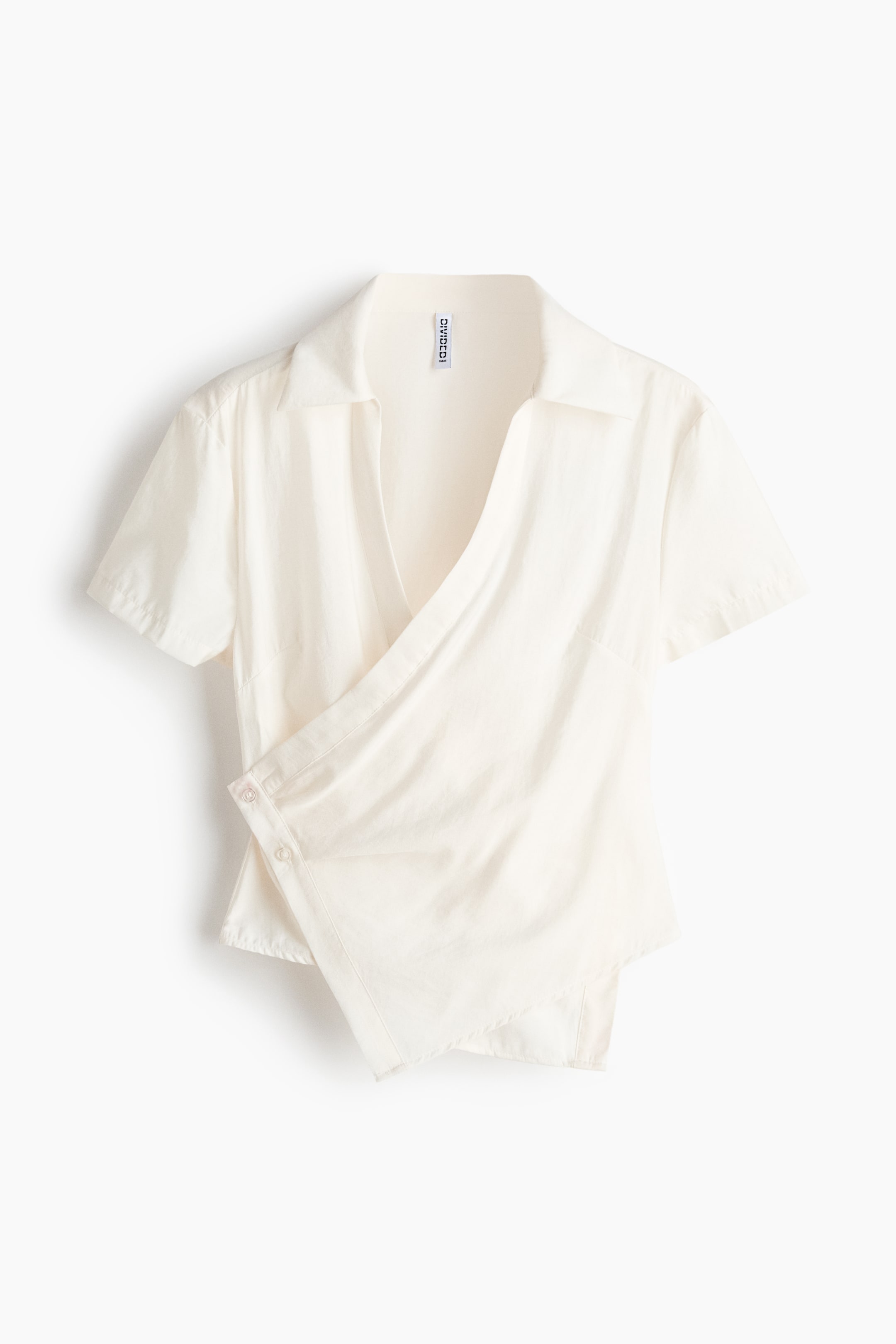 View larger image: A cream short-sleeved shirt featuring an open spread collar and an asymmetrical wrap front. The wrap is secured by two visible buttons on the lower left side.