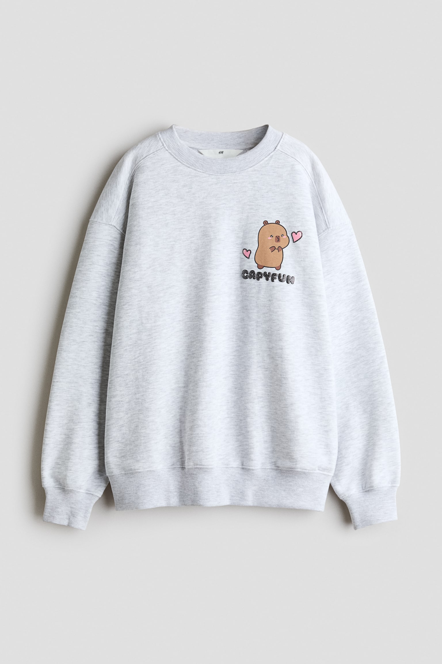 A light grey melange loose-fit sweatshirt is shown from the front, featuring a round rib-trimmed neckline, dropped shoulders, and ribbed cuffs and hem. The left chest displays a small graphic print of a cartoon capybara flanked by pink hearts, with the word "CAPYFUN" printed below.