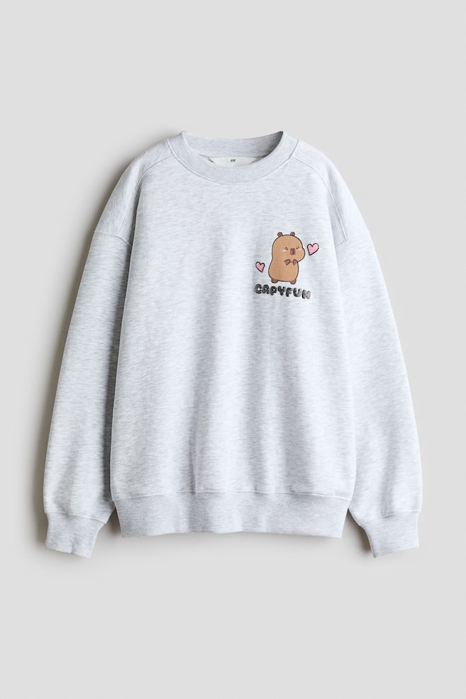 Un sweat-shirt ample chiné gris clair est présenté de face, avec un col rond bord-côte, des épaules tombantes, ainsi que des poignets et un ourlet côtelés. Sur la poitrine gauche figure un petit imprimé graphique représentant un capybara de style cartoon flanqué de cÅurs roses, avec le mot « CAPYFUN » imprimé en dessous. - 1