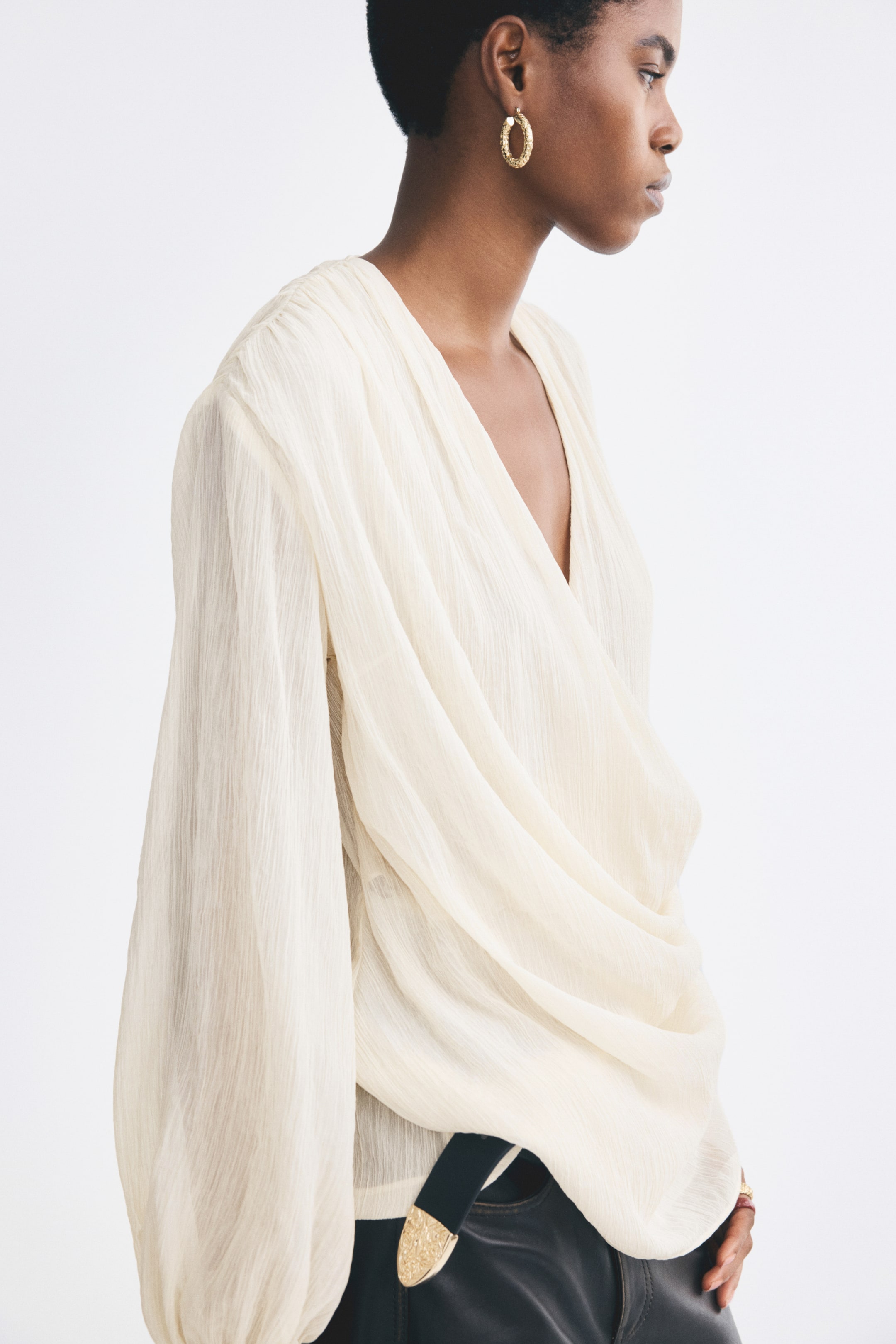 Visualizza immagine più grande: Una camicia color crema in tessuto stropicciato è indossata dalla modella, caratterizzata da una profonda scollatura a V, un design avvolgente drappeggiato sul davanti e maniche lunghe a palloncino. La camicia è abbinata a pantaloni scuri, infilata dentro, con una cintura nera e dorata.
