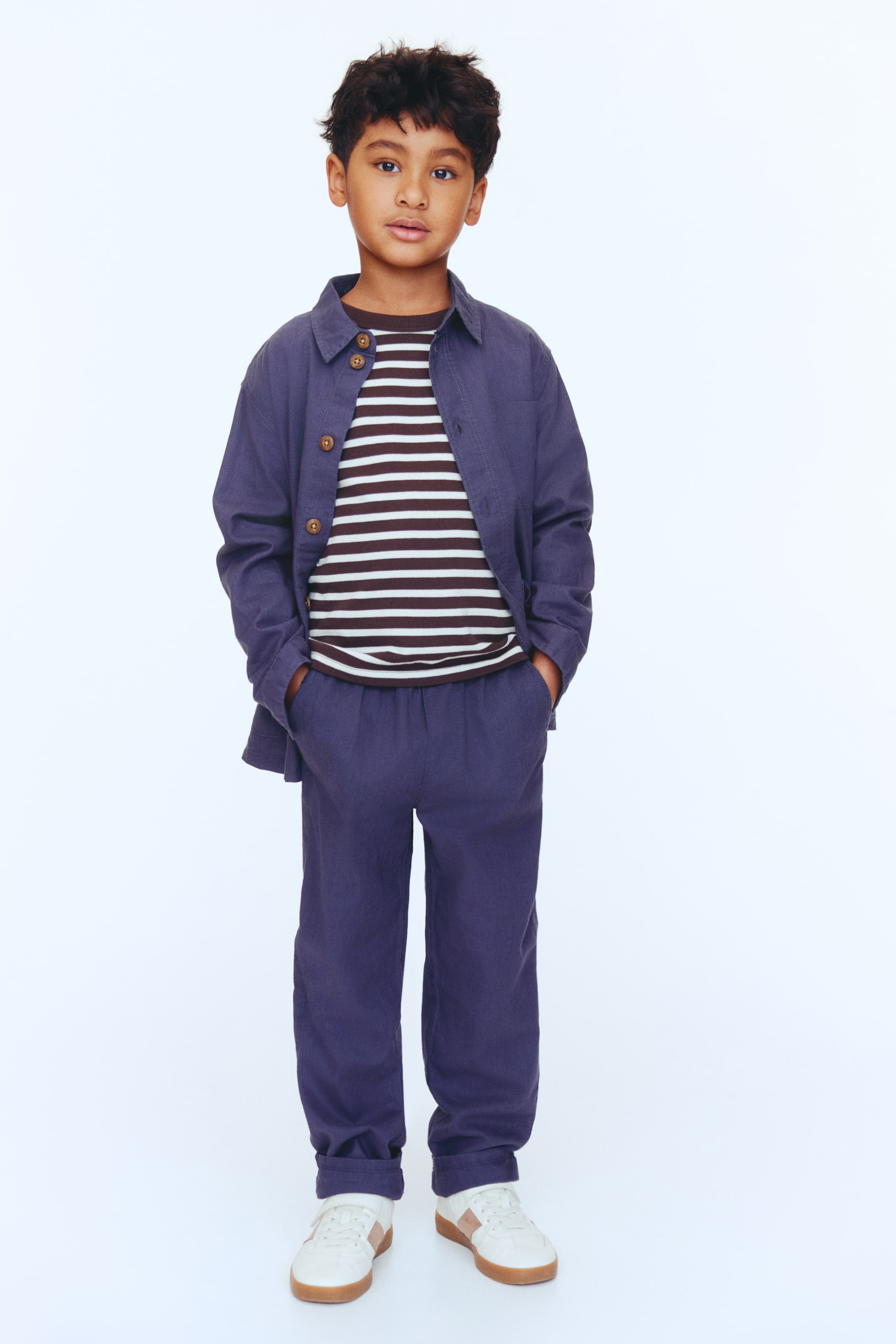 View larger image: Presenting a children's outfit that includes a dark blue collared shirt jacket, worn open over a brown and white striped t-shirt. The look is completed with matching dark blue pants, cuffed at the ankles, and white sneakers featuring brown accents and gum soles.