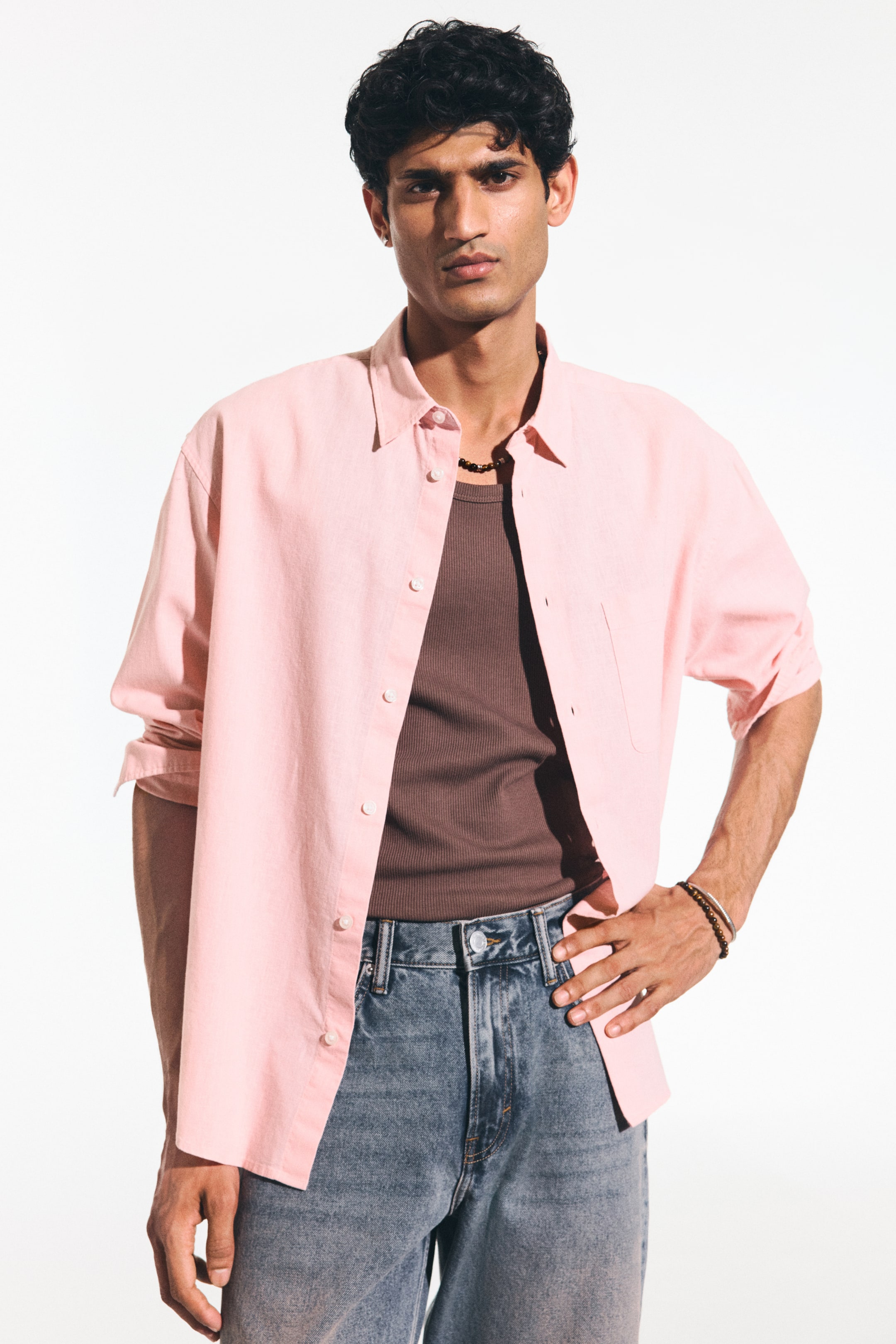View larger image: A pink button-down shirt featuring a pointed collar, white buttons down the front, and a patch pocket on the left chest. It is styled open over a brown ribbed tank top and partially tucked into blue jeans, with its long sleeves rolled up.