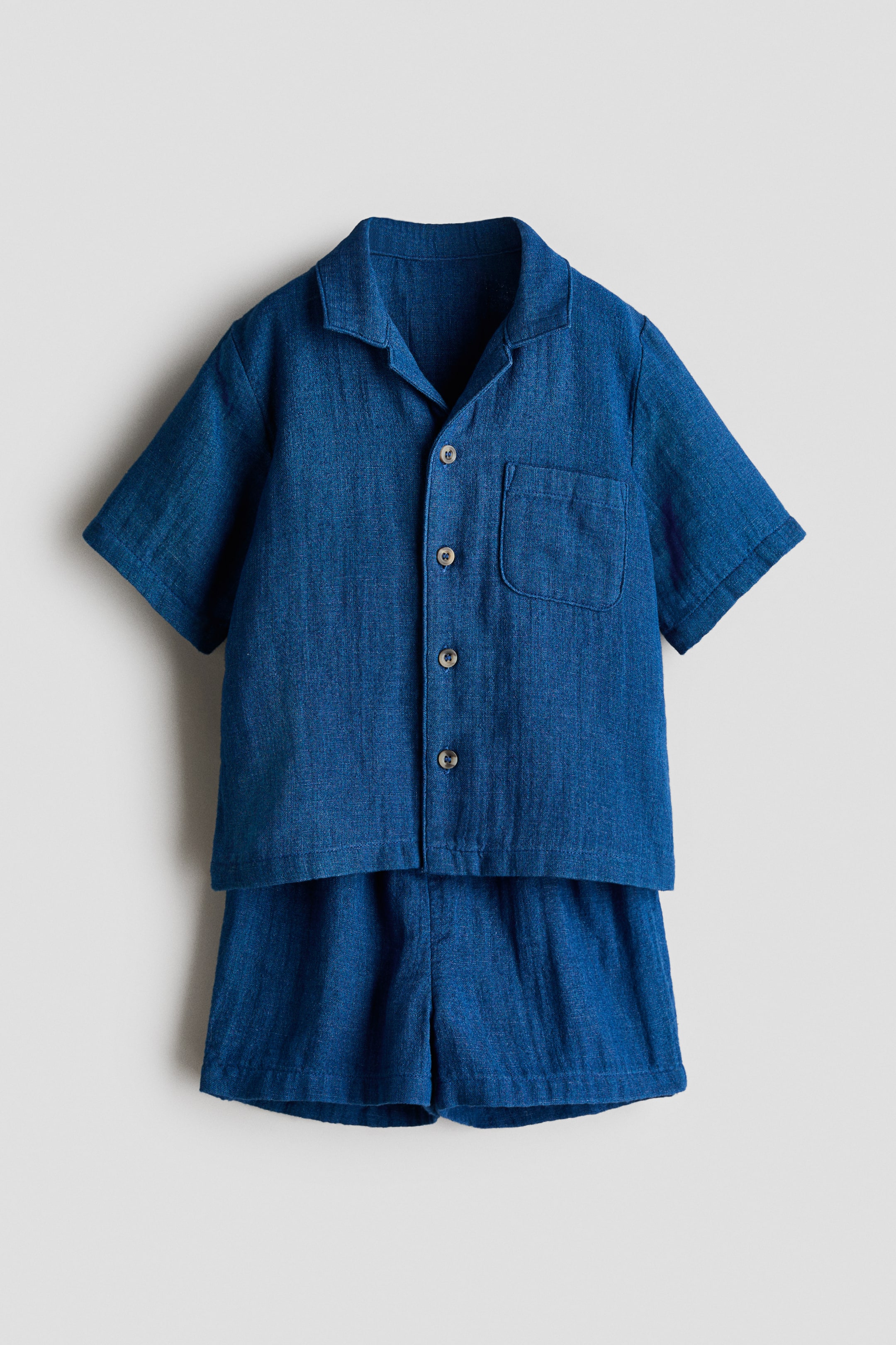 View larger image: Presenting a blue woven fabric ensemble, this set includes a straight-cut shirt paired with matching shorts, both showcased front-facing. The shirt features a resort collar, light-coloured buttons running down the front, short sleeves, and an open chest pocket. Meanwhile, the shorts exhibit a gathered waist.