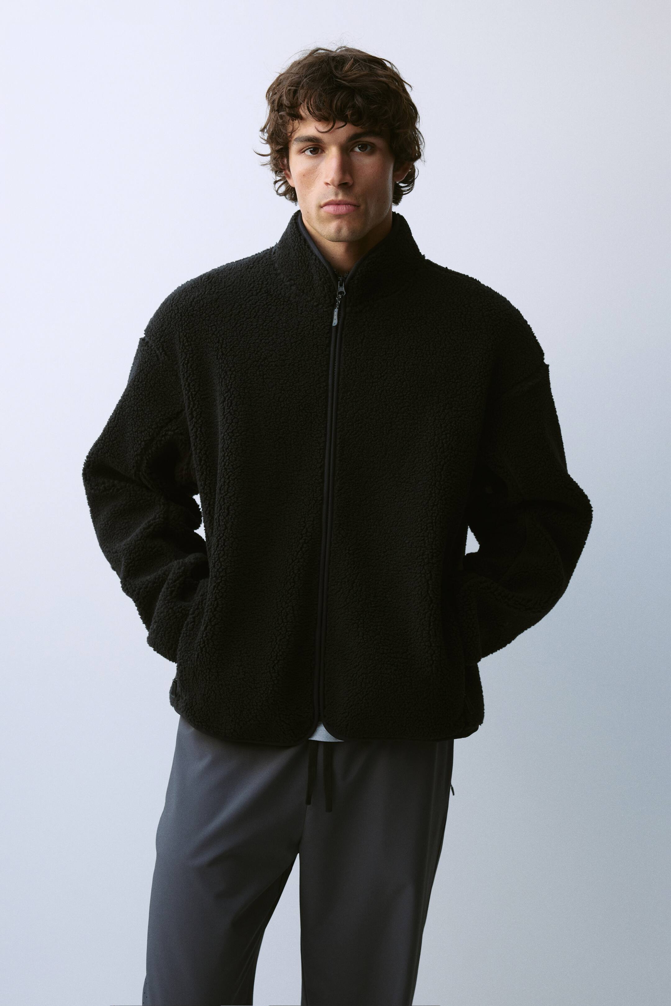 View larger image: A black teddy fleece activewear jacket featuring a relaxed fit, a stand collar, and a front zip closure is being worn by the model. The model is standing with hands placed inside the side welt pockets, pairing the jacket with dark grey trousers.