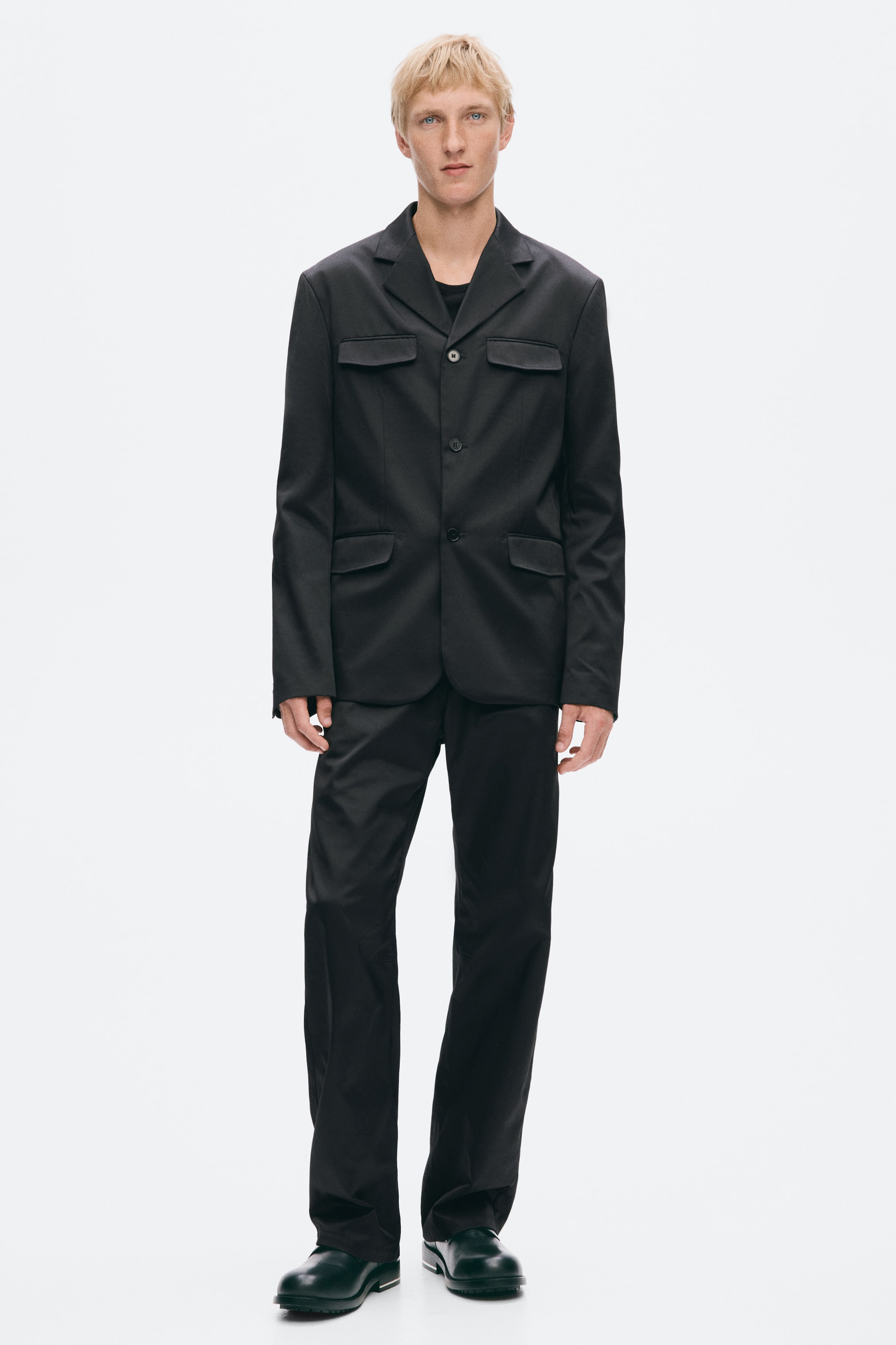 View larger image: The model is wearing a black single-breasted suit, featuring a structured blazer with notched lapels, three front buttons, two chest flap pockets and two hip flap pockets. The suit is styled with matching straight-leg trousers, a black crew neck T-shirt and dark leather shoes.