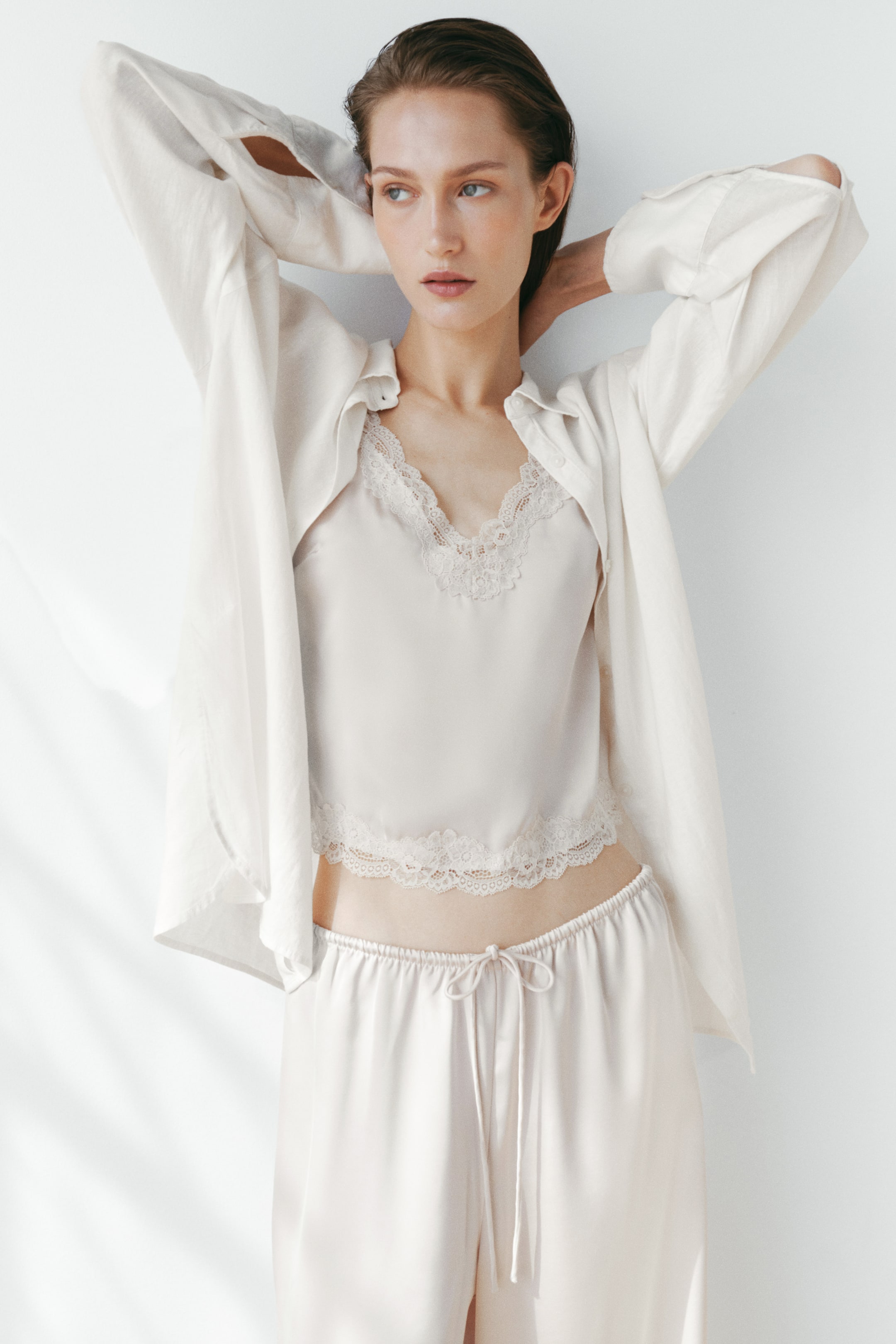 View larger image: An oversized light-coloured shirt in an airy weave, featuring a collar, dropped shoulders, and long buttoned sleeves, is being worn open over a lace-trimmed camisole. The model is raising her arms, causing the shirt to drape around her.