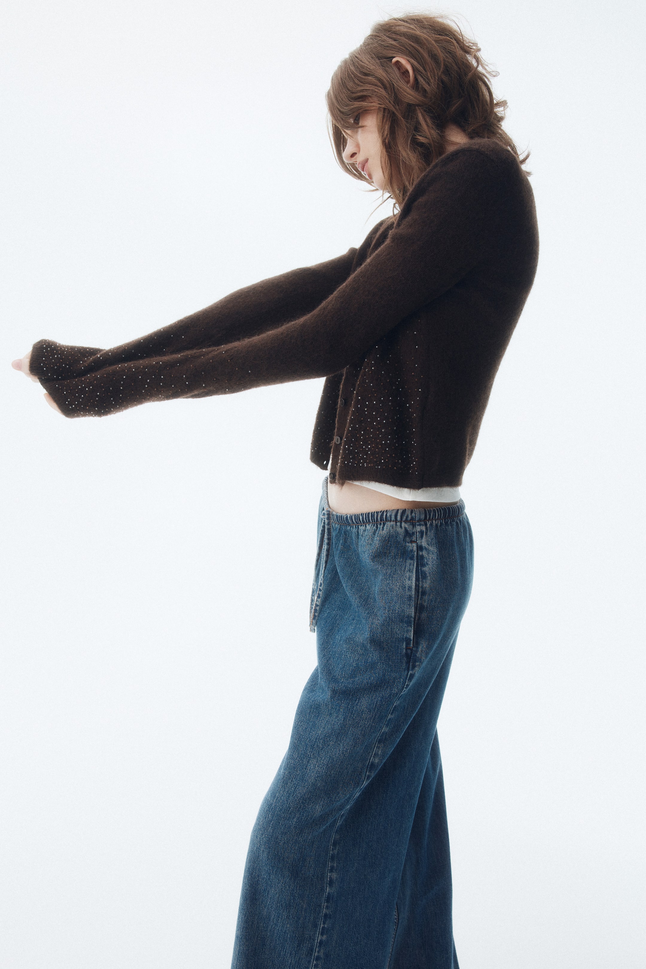 Ampliar la imagen: Un cárdigan corto de color marrón se ve desde un ángulo lateral, con sus brazos extendidos hacia adelante, mostrando pequeños adornos de brillantes diseminados por el frente y las mangas. Se lleva sobre una camiseta blanca y se combina con jeans de pierna ancha azules con una cintura elástica con cordón.