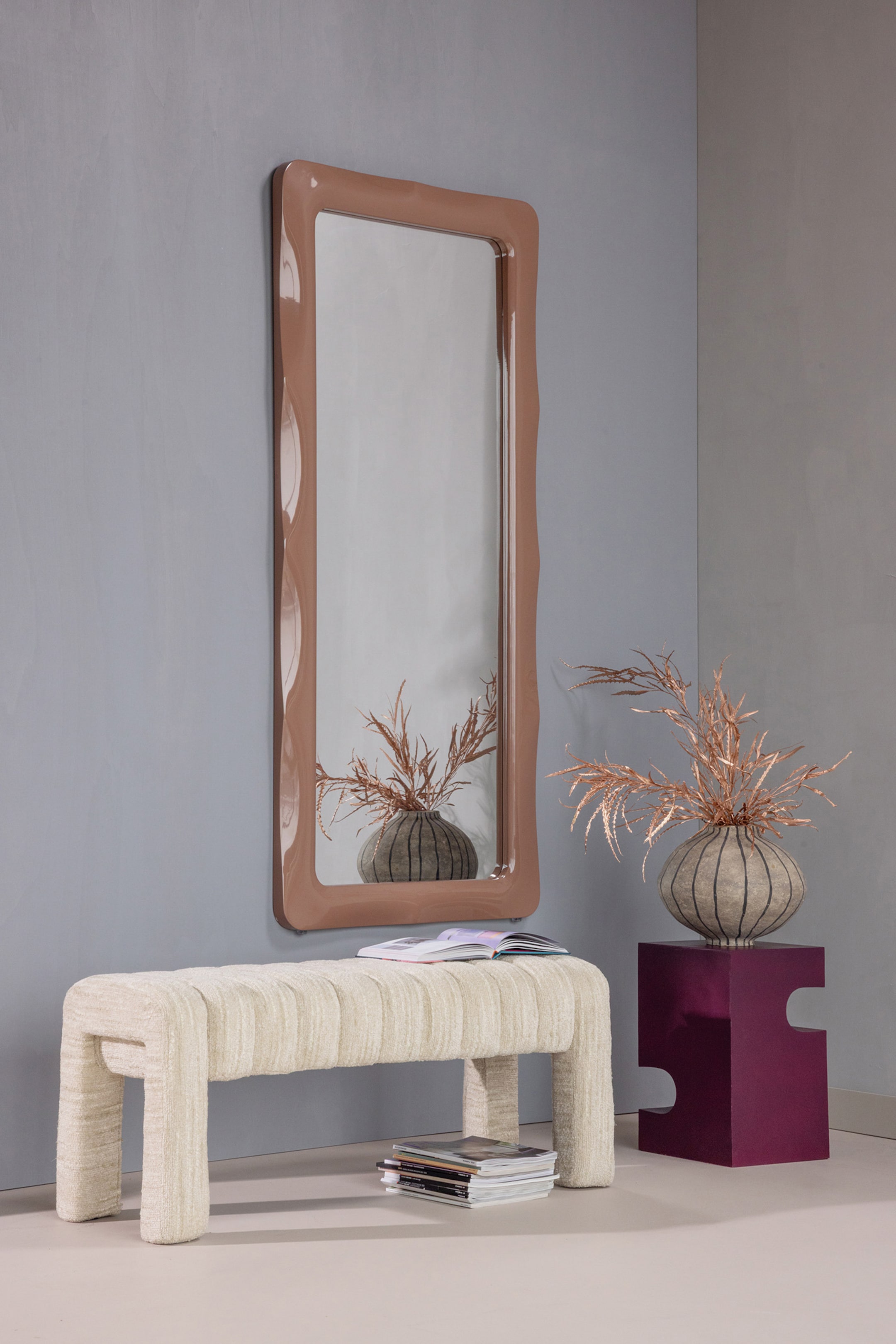 Agrandir l'image: Un grand miroir rectangulaire pleine longueur arbore un cadre ondulé, brillant et de couleur taupe chaud, reflétant un vase décoratif avec des herbes séchées et un banc texturé.