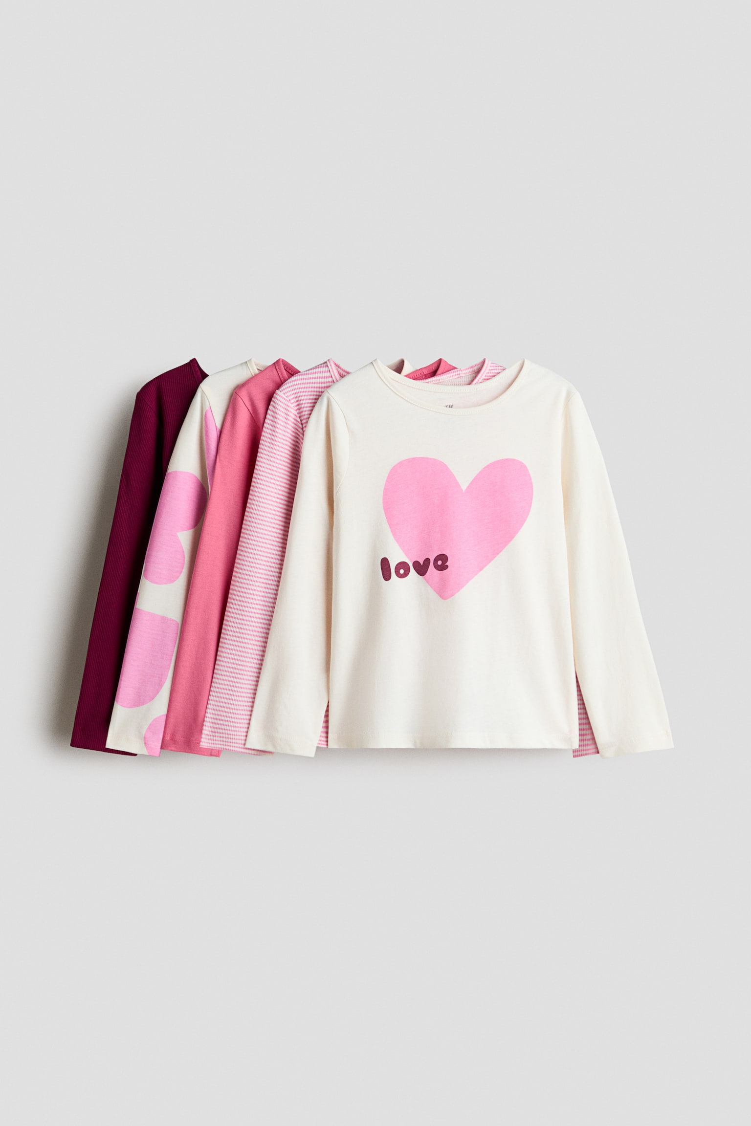 Five long-sleeve cotton jersey t-shirts are overlapping in a row facing forward, featuring round necklines with narrow trim. The front cream-colored shirt is displaying a large pink heart graphic with the word "love" printed on it, while the shirts layered behind are showing pink-and-white stripes, solid pink, pink heart patterns, and solid dark burgundy.