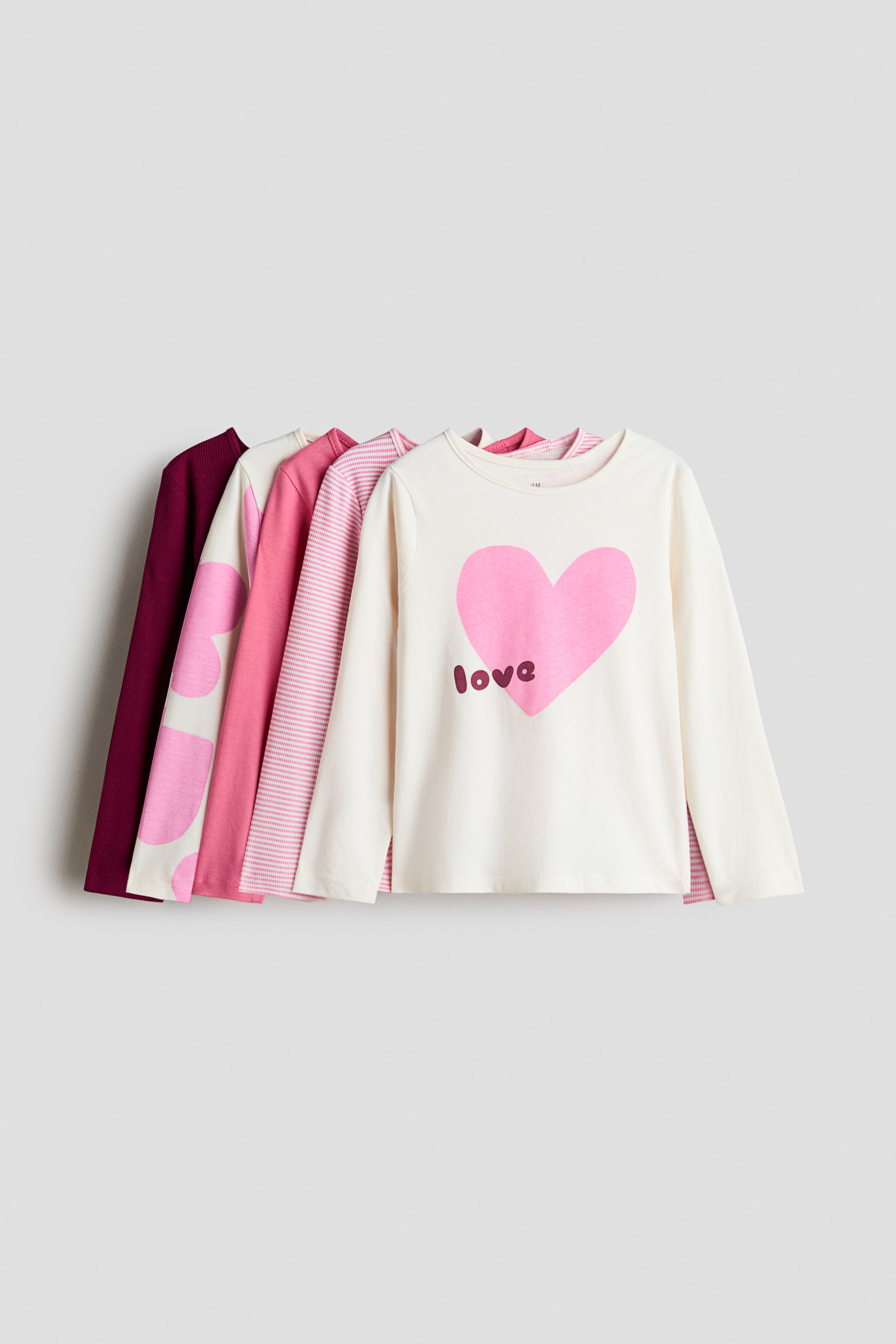 Grotere afbeelding bekijken: Vijf T-shirts van katoenen jersey met lange mouwen liggen in een rij overlappend en zijn van voren weergegeven, met ronde halslijnen met een smalle afwerking. Het voorste T-shirt in crèmekleur toont een grote roze hartafbeelding met het woord "love" erop gedrukt, terwijl de shirts daarachter roze-witte strepen, effen roze, roze hartpatronen en effen donker bordeaux laten zien.