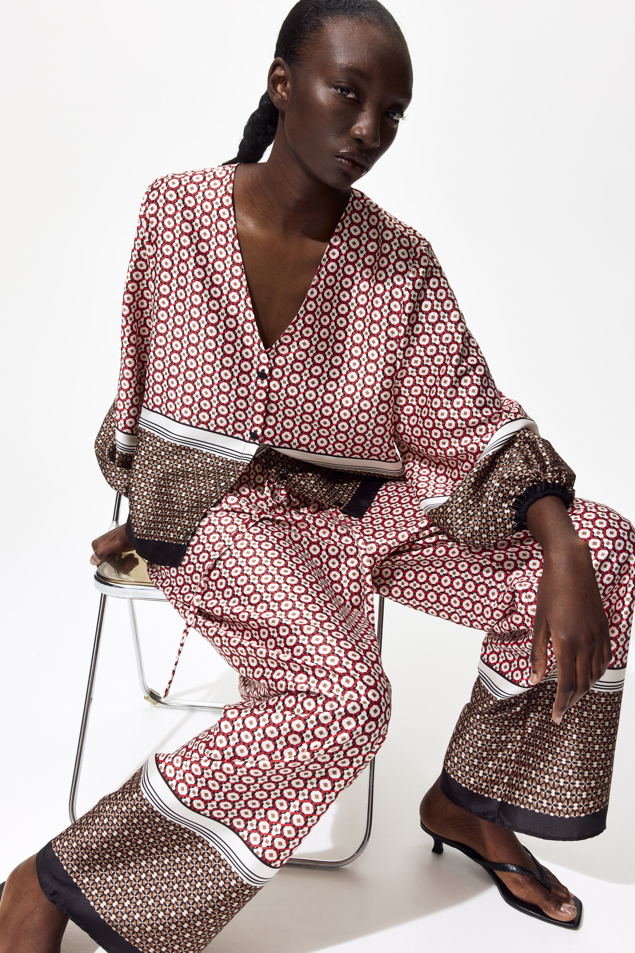 View larger image: The model is wearing loose-fit trousers crafted from woven fabric with a subtle sheen. These trousers boast an elasticated, drawstring waist, straight legs, and are adorned with red, white, and black geometric patterns, complemented by a contrasting geometric band near the hem.