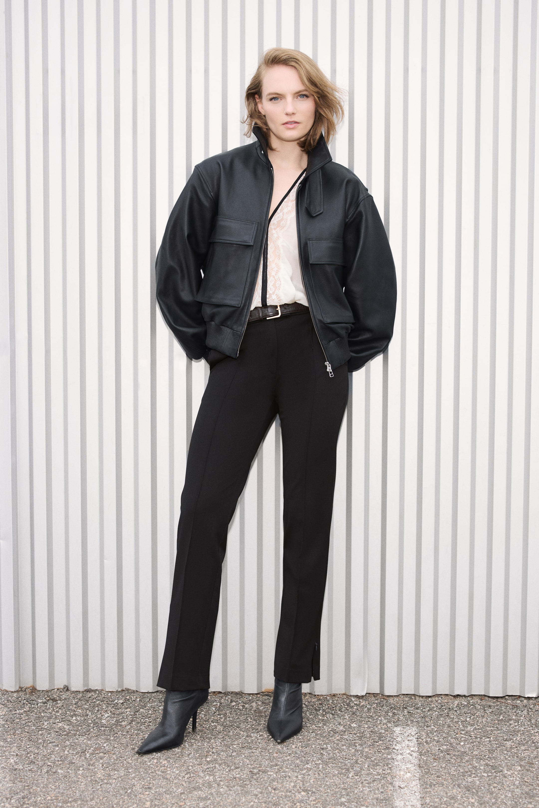 Visualizza l'immagine ingrandita: Pantaloni neri sartoriali con gamba slim, pieghe frontali ben definite e cintura scura in vita, indossati dalla modella. I pantaloni sono abbinati a un bomber in pelle nera, una blusa in pizzo color crema e stivali neri con tacco e punta affusolata.