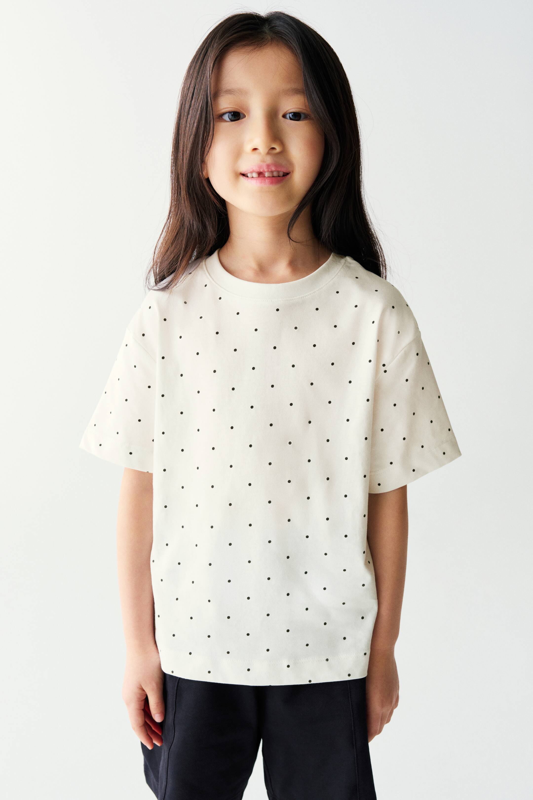 View larger image: An off-white, short-sleeved t-shirt featuring a pattern of small black polka dots is being worn by the model. The t-shirt has a crew neckline and a relaxed fit with dropped shoulders, and is styled with black pants.