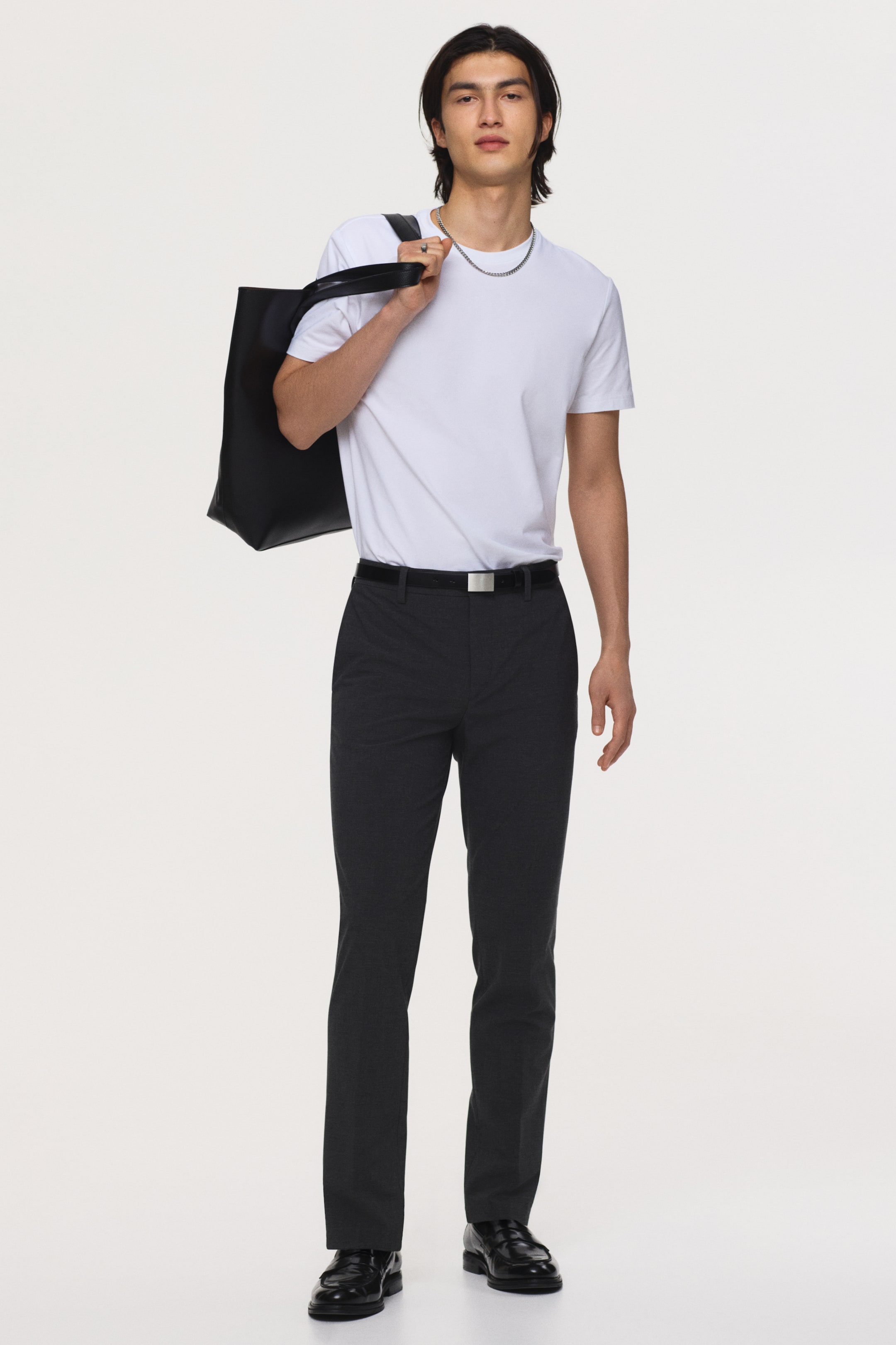 Visa större bild: Mörkgrå slim-fit byxor bärs, med ett subtilt texturerat mönster, raka ben och en synlig framveck. De hålls uppe av ett svart bälte med ett silverfärgat spänne, och en vit t-shirt är instoppad i dem.