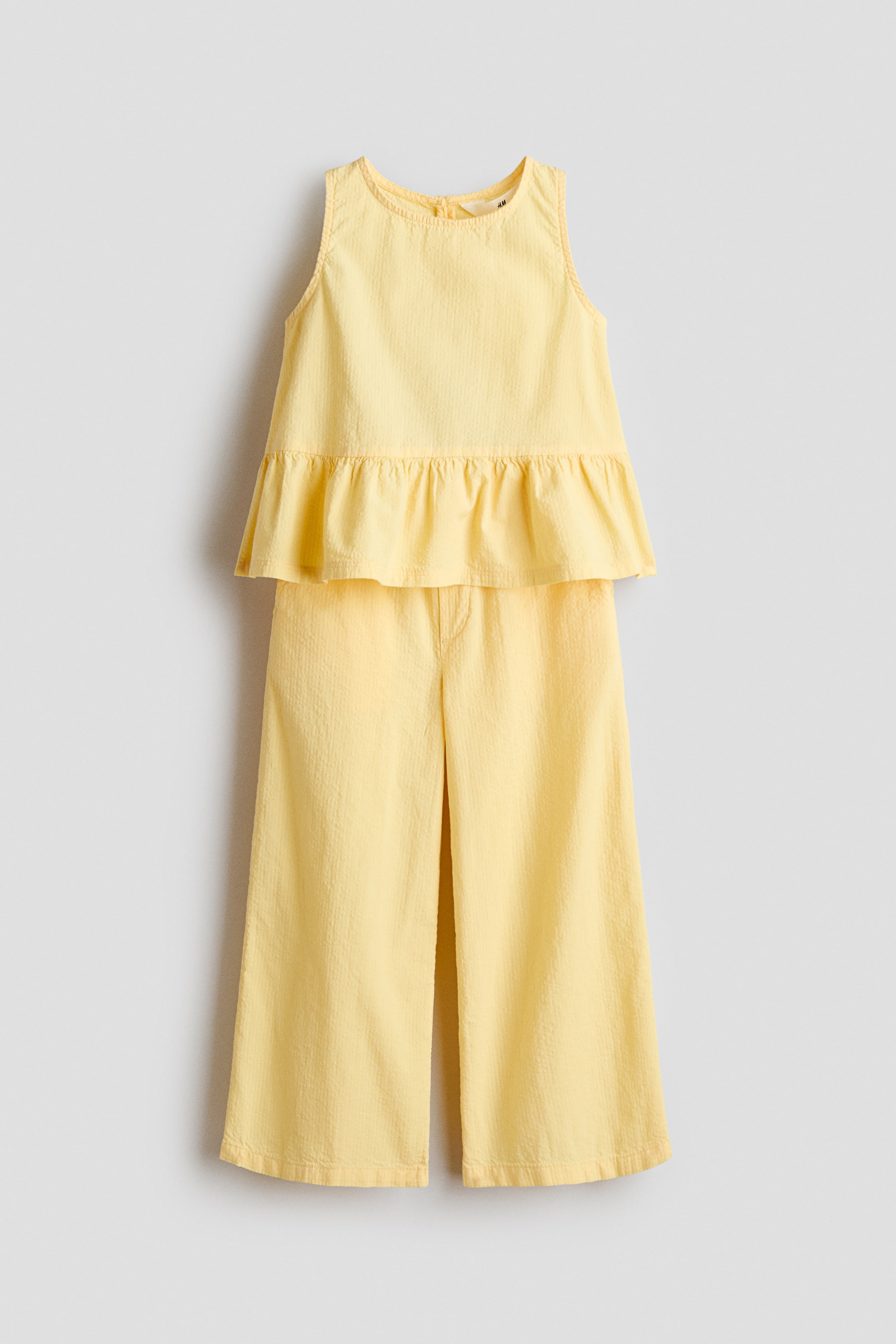 View larger image: A yellow two-piece outfit is shown from the FRONT. The sleeveless top features a round neckline and a ruffled peplum hem. The wide-leg trousers are made from textured fabric and have a cropped length.