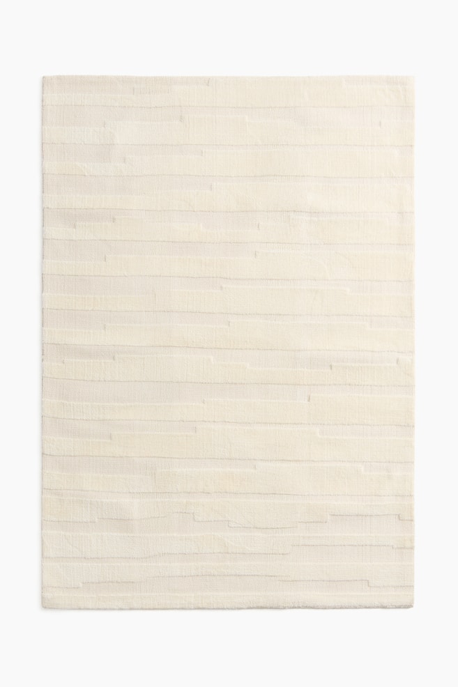 Una alfombra tejida presenta un patrón texturizado, vista desde el frente. Cuenta con bandas horizontales de alturas de pelo y texturas variables, creando un diseño de rayas irregulares. - 1