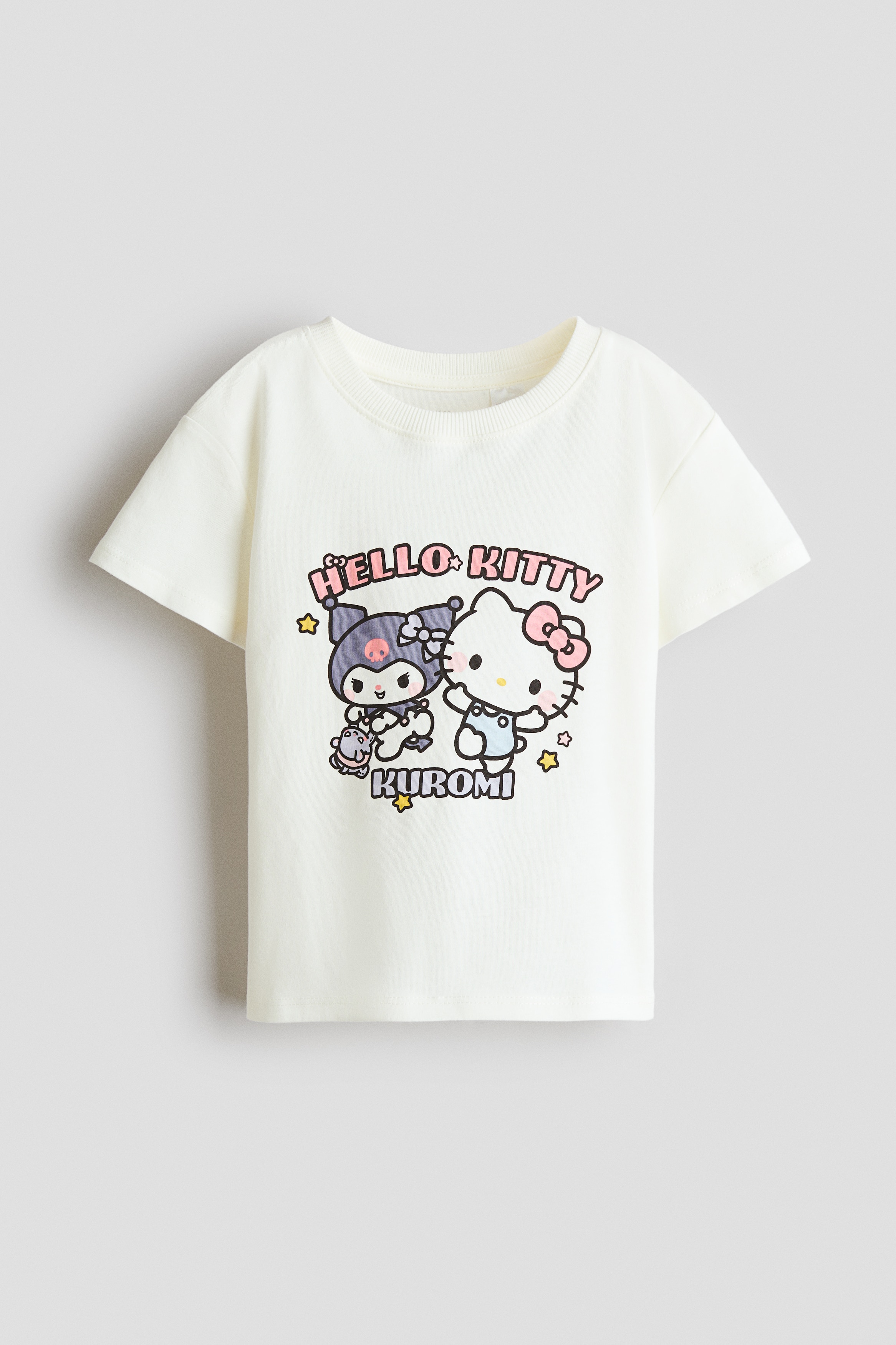 A cream-coloured cotton jersey T-shirt featuring a graphic print of Hello Kitty and Kuromi on the front is worn by the model. The T-shirt is designed with a round, rib-trimmed neckline and dropped shoulders, and is paired with blue denim pants.