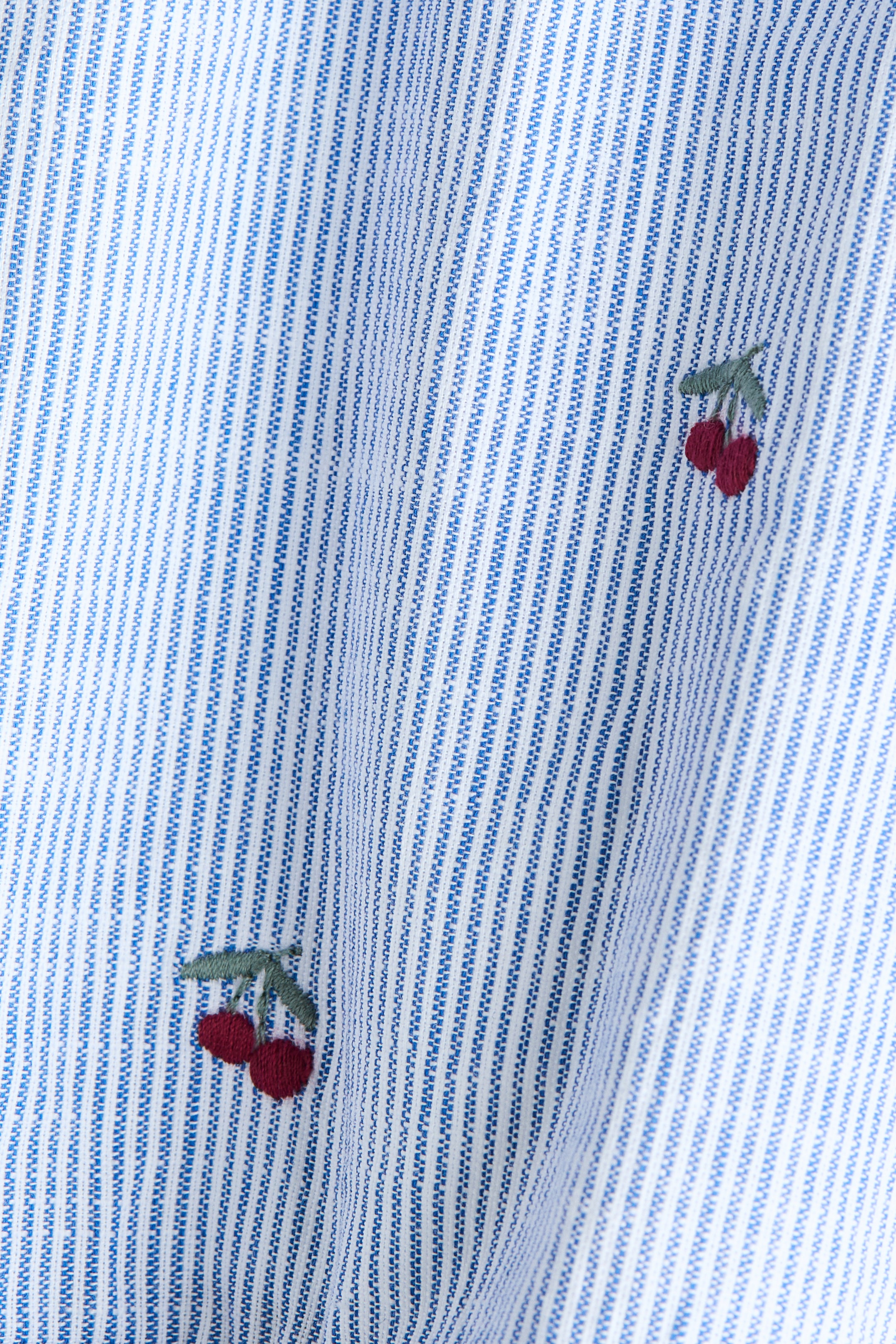 View larger image: The fabric showcases alternating vertical stripes in hues of blue and white, highlighted by its textured weave. Scattered across the gently flowing material are small embroidered cherry motifs, each depicting a pair of dark red cherries with a green stem.