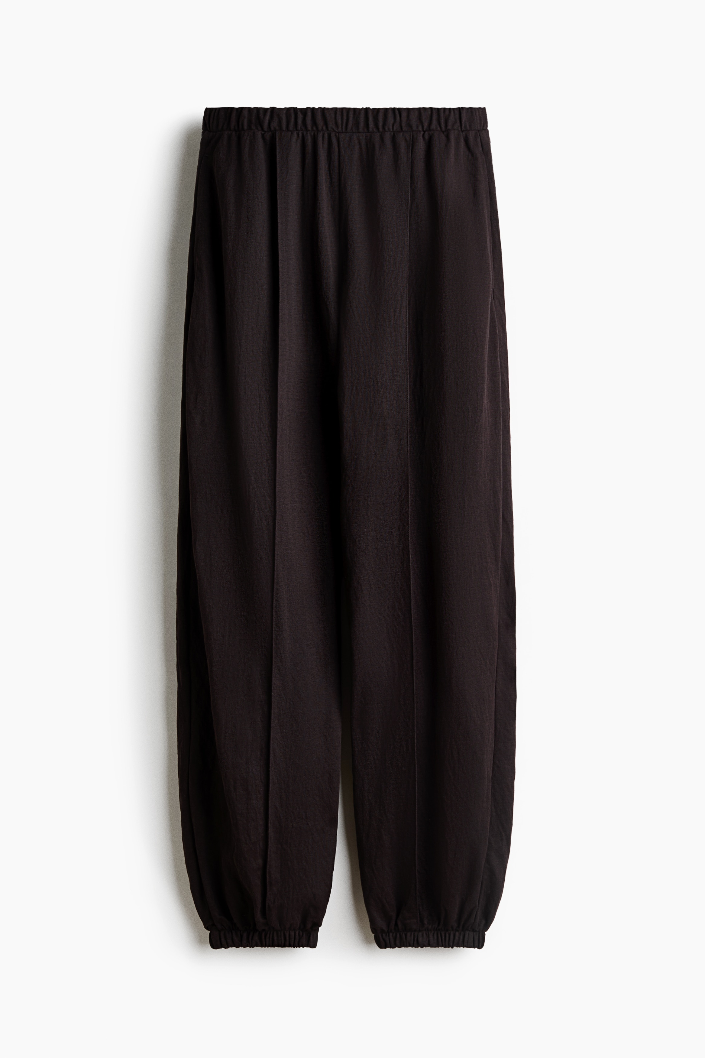 La modelo lleva puestos unos pantalones globo de jersey color cafÃ© oscuro y corte holgado, mientras coloca una mano dentro del bolsillo lateral. Los pantalones, a la altura del tobillo, cuentan con dobladillos fruncidos con elÃ¡stico y se combinan con una blusa a juego de cuello alto y sandalias negras de tacÃ³n.