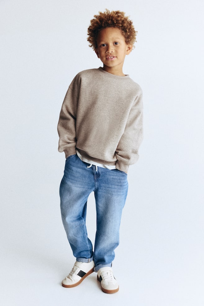 Bir çocuğun kıyafeti, rahat kesimli, nervürlü manşetleri ve nervürlü eteği olan açık kahverengi bir bisiklet yaka sweatshirt'ü, beyaz bir tişört üzerine katmanlı olarak ön plana çıkarıyor. Sweatshirt'ü tamamlayan açık yıkamalı mavi kot pantolonlar, bağcık bel ve manşetli bilekler gösterirken, koyu yan şeritler ve kahverengi kauçuk tabanlı beyaz düşük-top spor ayakkabılar kombini tamamlıyor. - 1