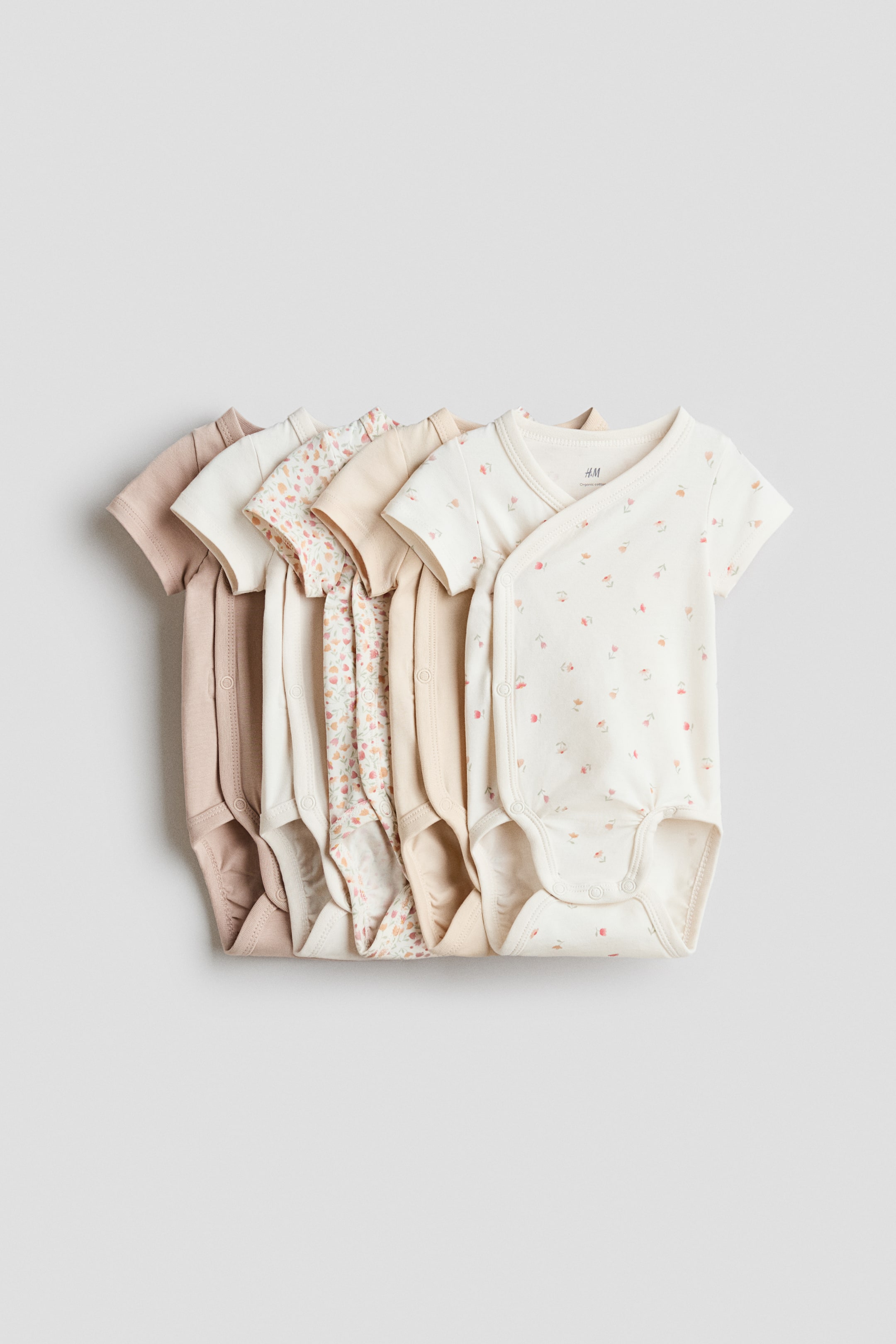 View larger image: A set of short-sleeve bodysuits in soft cotton jersey featuring wrapover fronts and snap closures at the sides and crotch, shown from the FRONT. The layered bodysuits come in solid beige, cream, and floral patterns.