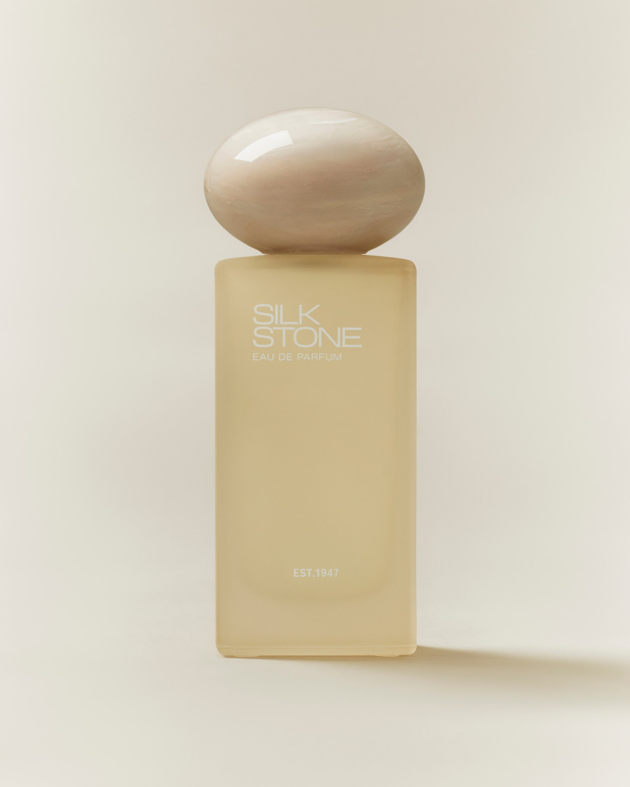 Ver imagen más grande: Un frasco de perfume presenta un cuerpo rectangular, de un suave color amarillo pálido esmerilado, coronado con un tapón esférico de mármol crema. El frasco está orientado hacia adelante, mostrando "SILK STONE EAU DE PARFUM" y "EST.1947" en texto blanco en su parte frontal.