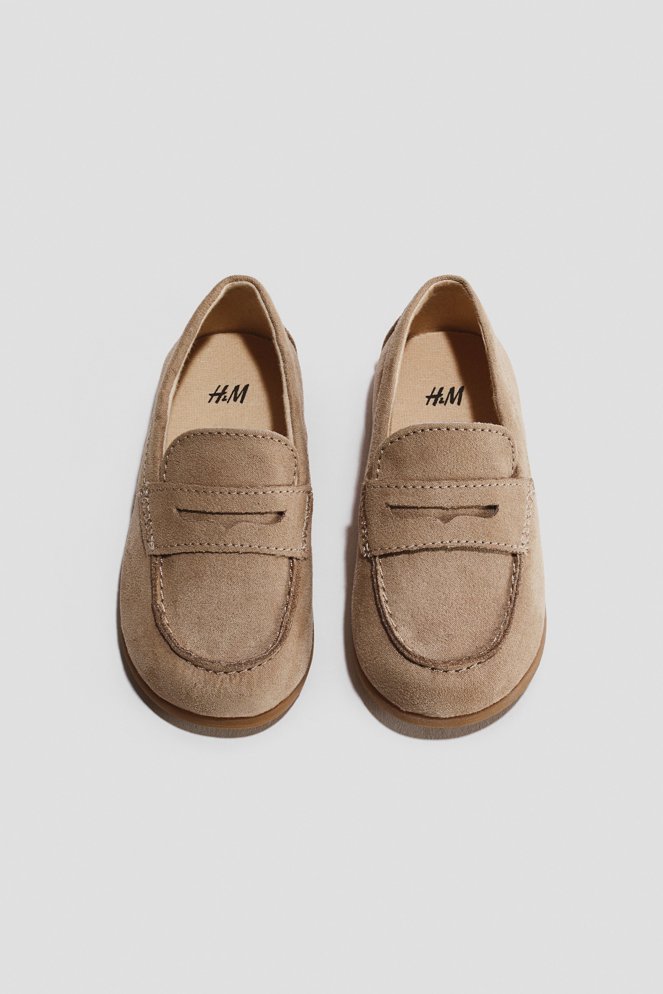 View larger image: A pair of light brown faux suede baby loafers featuring a classic penny strap across the vamp and moc-toe stitching on the toe box. The H&M logo is visible on the light-coloured interior lining of each shoe.