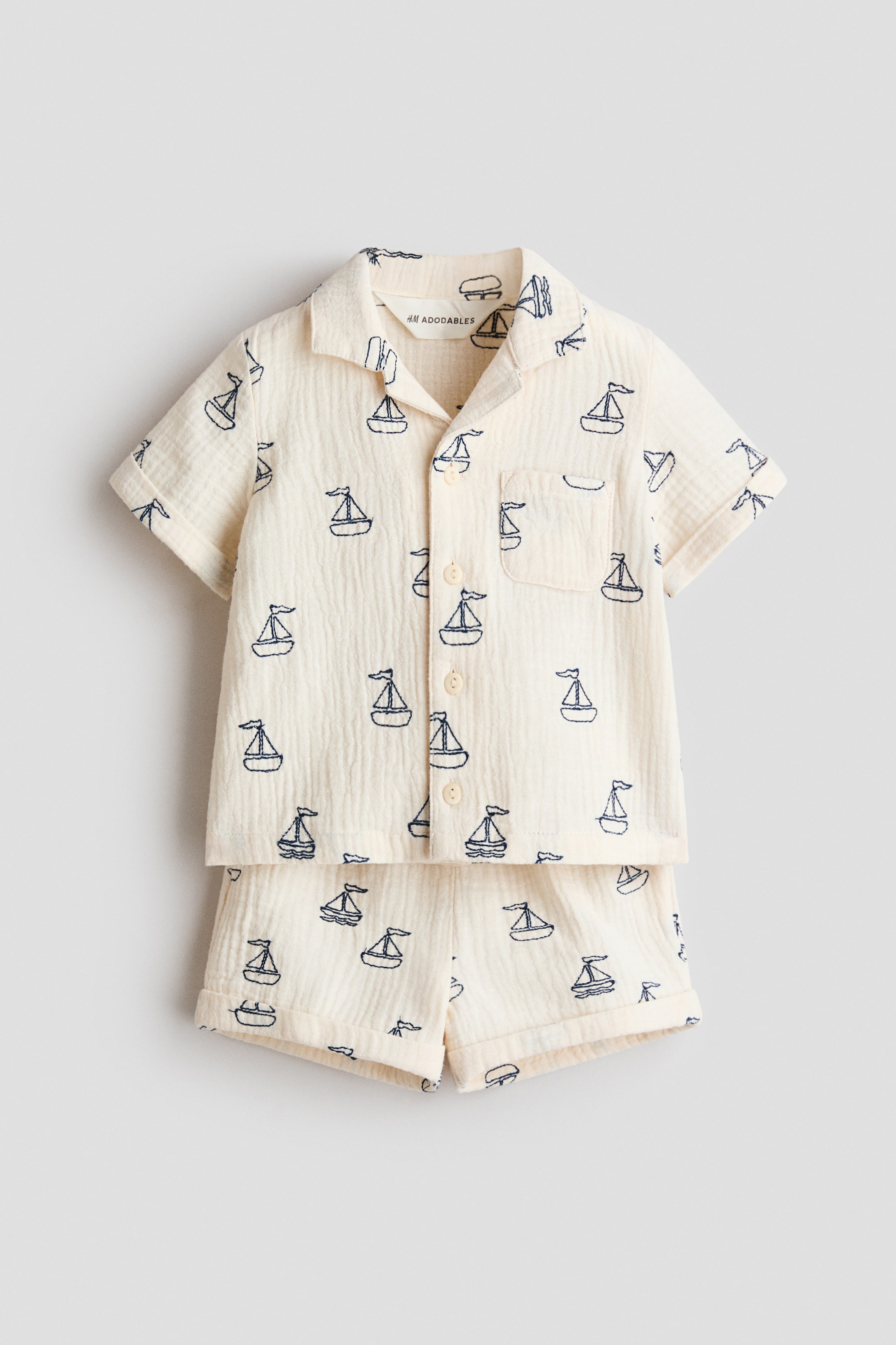 Voir une image plus grande: Un ensemble deux pièces crème, vu de face, comprenant une chemise à manches courtes et un short assorti, tous deux arborant un motif de voilier bleu foncé sur un tissu de gaze froissé. La chemise présente un col camp, des manches courtes avec revers, une patte de boutonnage frontale avec des boutons crème, et une poche poitrine unique, tandis que le short assorti affiche des ourlets à revers.