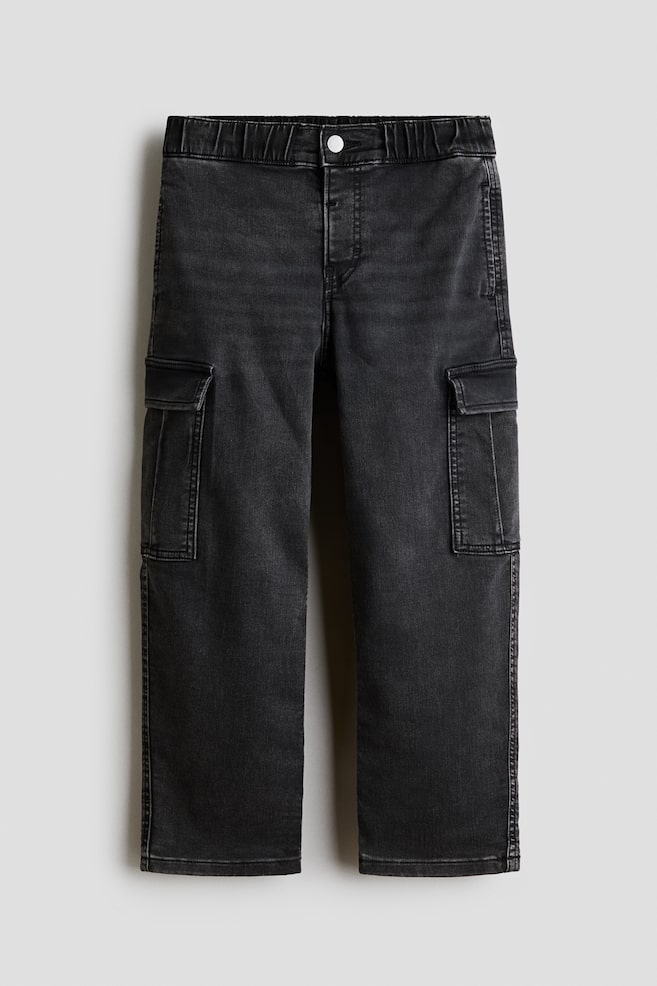 A pair of washed black denim cargo trousers is laid flat facing front, featuring an elasticated waistband with a single button fastening and a straight-leg cut. The design features two large utility cargo pockets with flap closures on the sides of the thighs. - 1