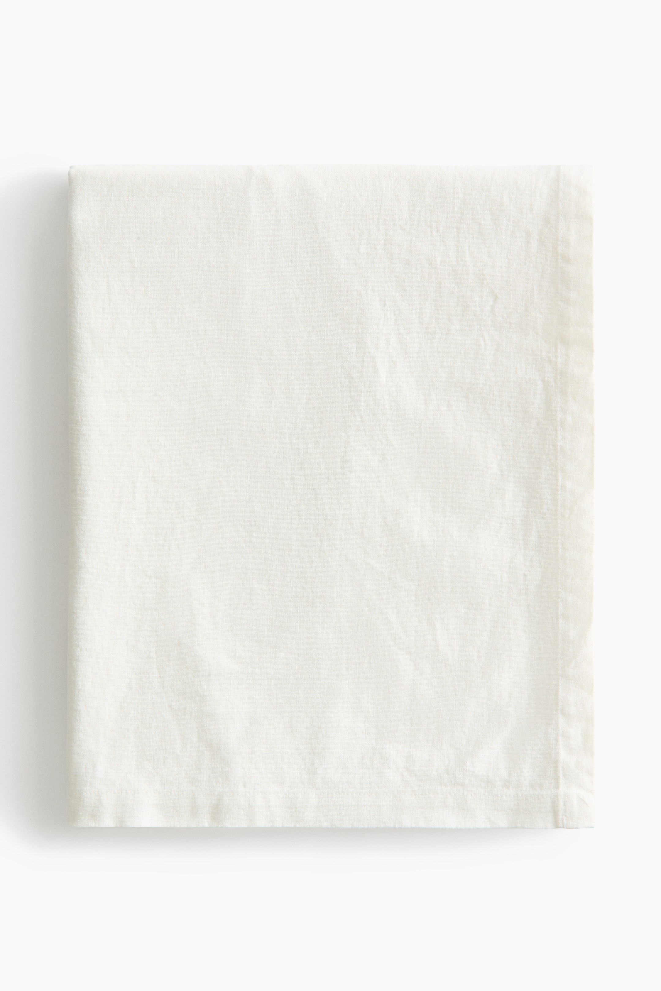 View larger image: A cream-colored tablecloth is laid out flat, neatly folded into a rectangle. It showcases a gentle woven texture with noticeable creases and features a defined folded hem along its edges.