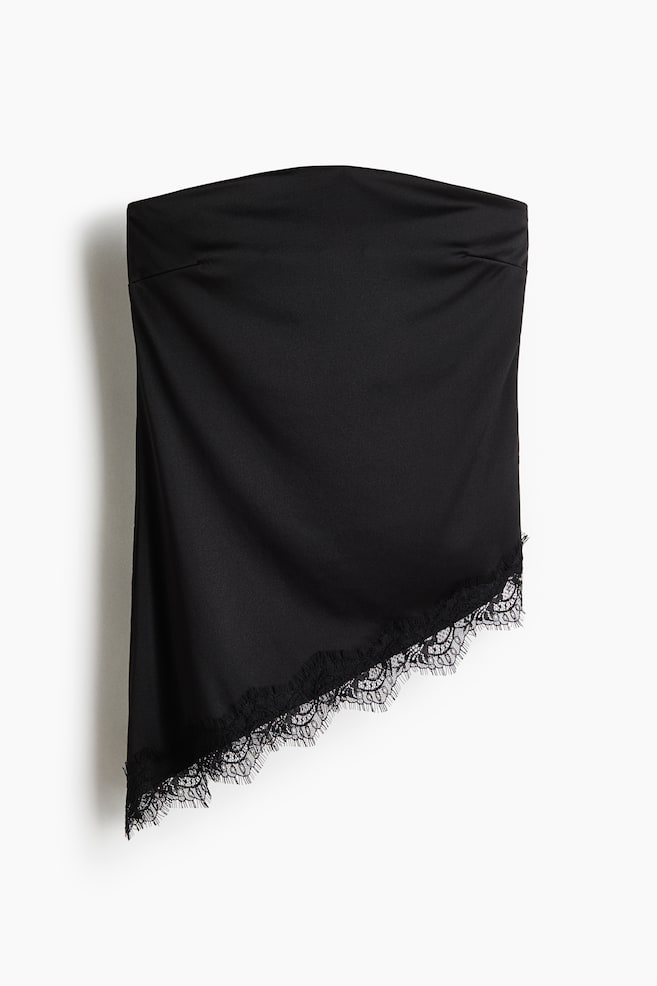 A black strapless top is displayed from the front, featuring smooth black fabric and a gathered band along its top edge. The hem is cut asymmetrically and is trimmed with delicate black lace and fringed detailing. - 1