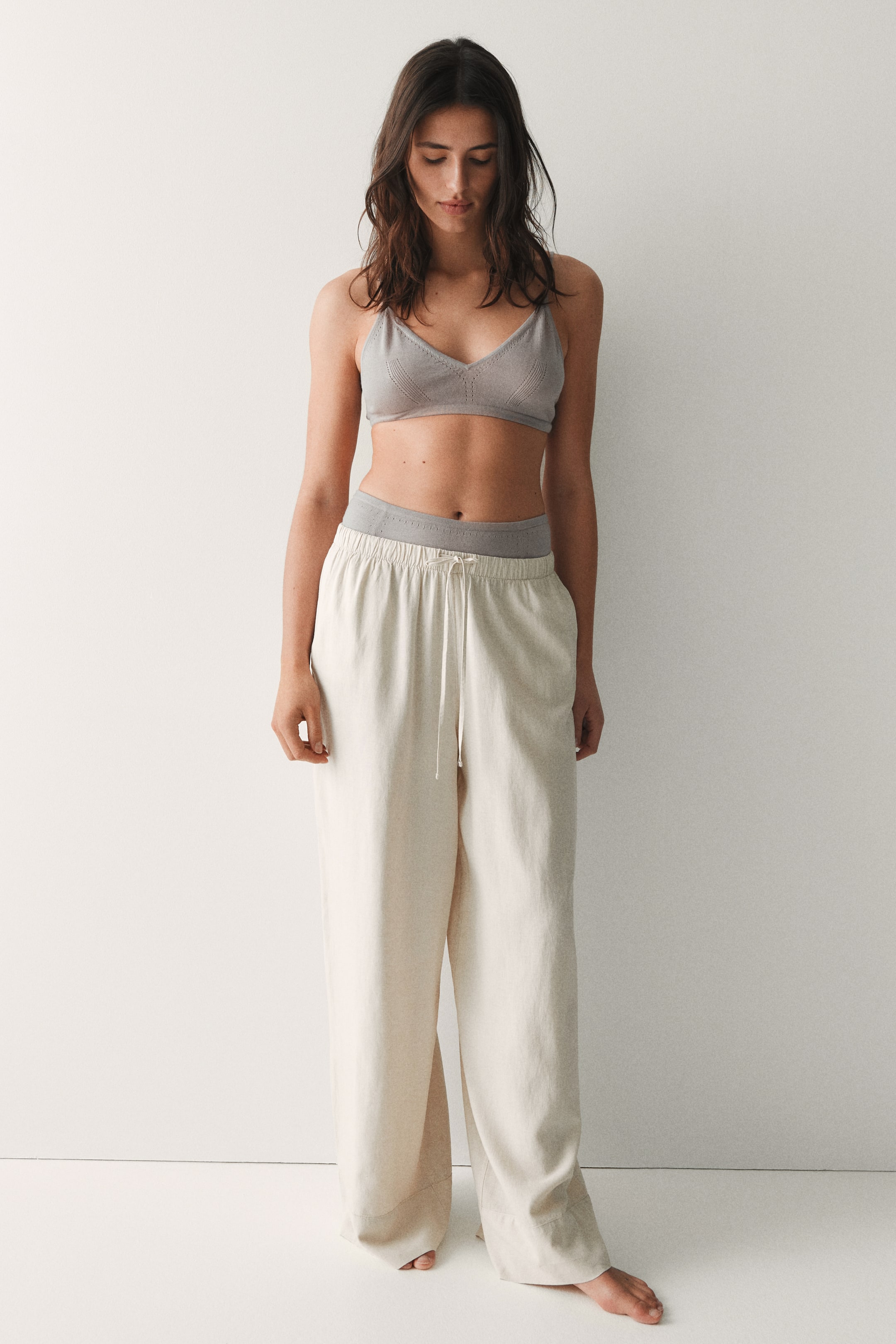 View larger image: A light grey knit bralette showcases triangle cups with subtle perforated details and a wide solid underband. Cream-coloured wide-leg pants are worn, featuring an elasticated drawstring waistband, with a grey undergarment band visible above the pants' waistline.