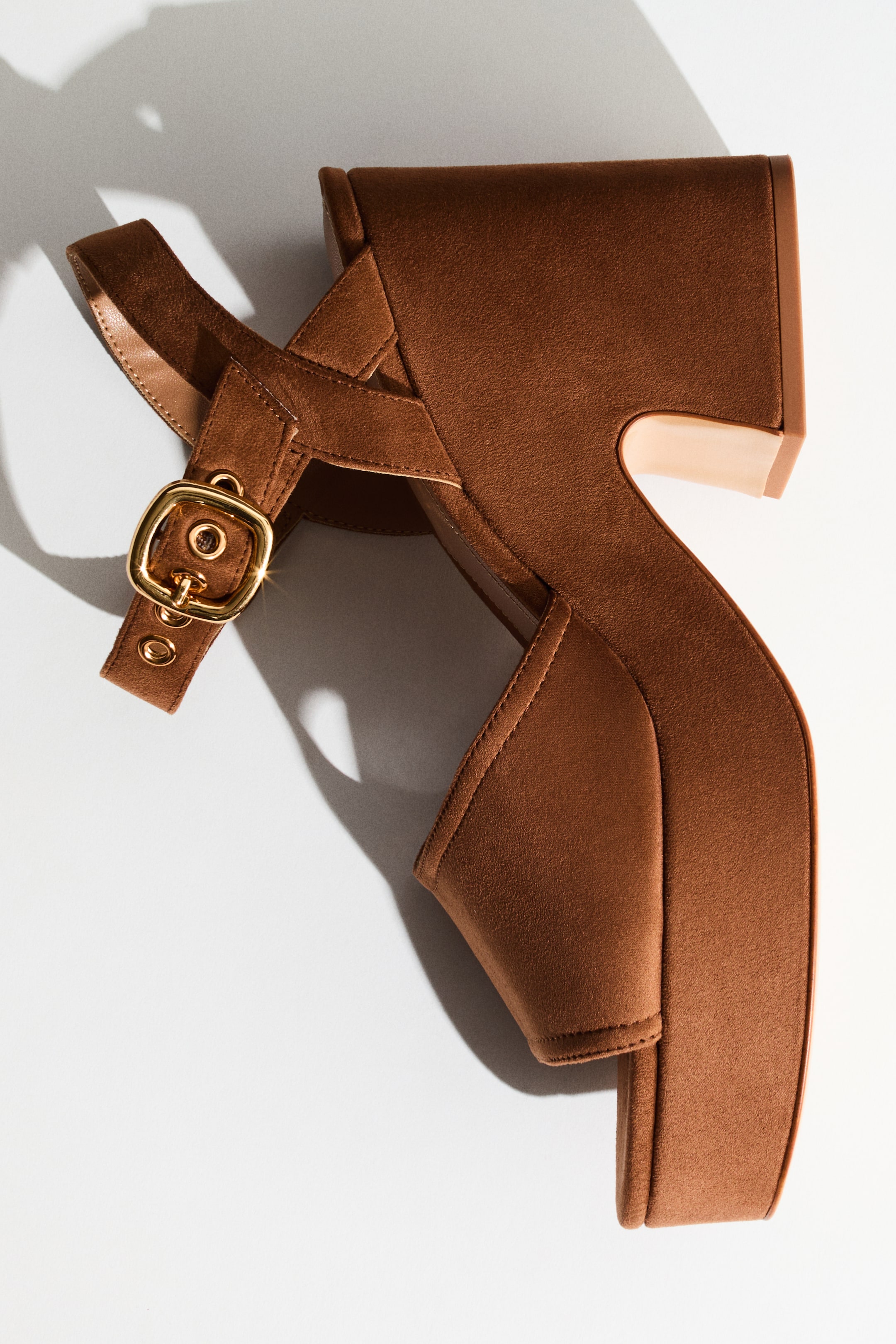 View larger image: A brown platform sandal crafted from a suede-like material is displayed at an angle, highlighting its side profile. It features a wide forefoot strap and an ankle strap with a gold-toned buckle, along with a chunky block heel that has an angular cutout detail, revealing a lighter inner surface.