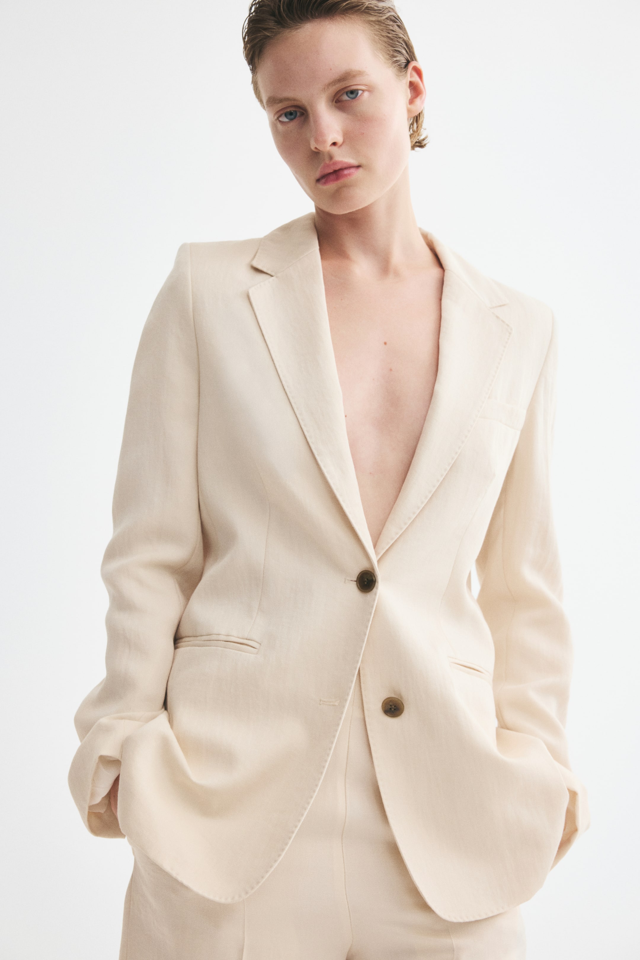 View larger image: A long, single-breasted blazer in viscose-blend twill is worn by the model, featuring notch lapels, a front button closure, waist darts, a welt chest pocket, and jetted front pockets where the model’s hands are resting. The blazer is styled without an undershirt and paired with matching pants.