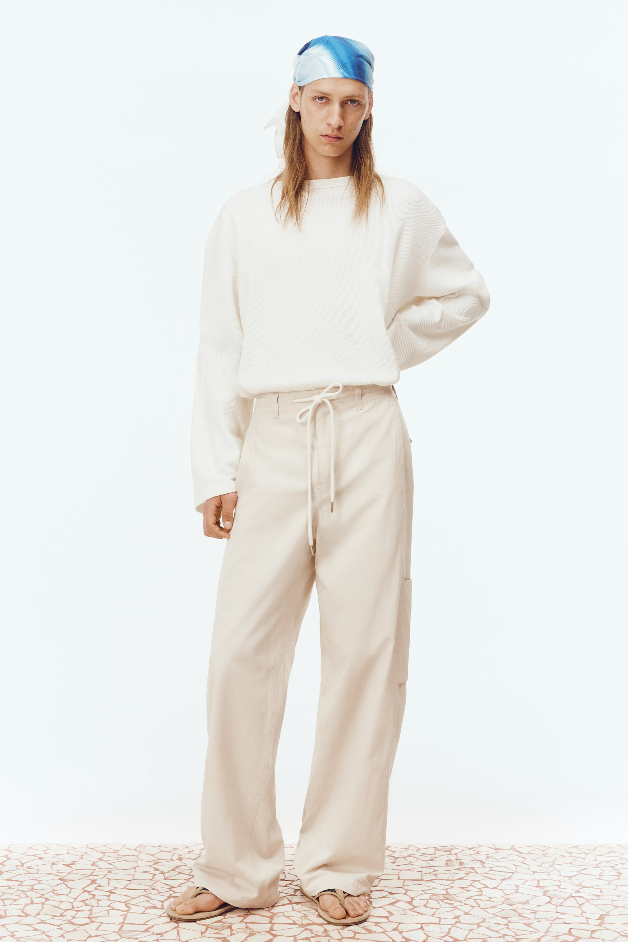Büyük resmi görüntüle: Model, krem rengi bir bisiklet yaka sweatshirt ve bej rengi geniş paça kargo pantolon giymektedir. Pantolonun belirgin bir kargo cebi ve bağcıklı beli vardır. Modelin başında mavi ve beyaz desenli bir başörtüsü bulunmaktadır ve ayaklarında açık renkli parmak arası sandaletler giyilmektedir.