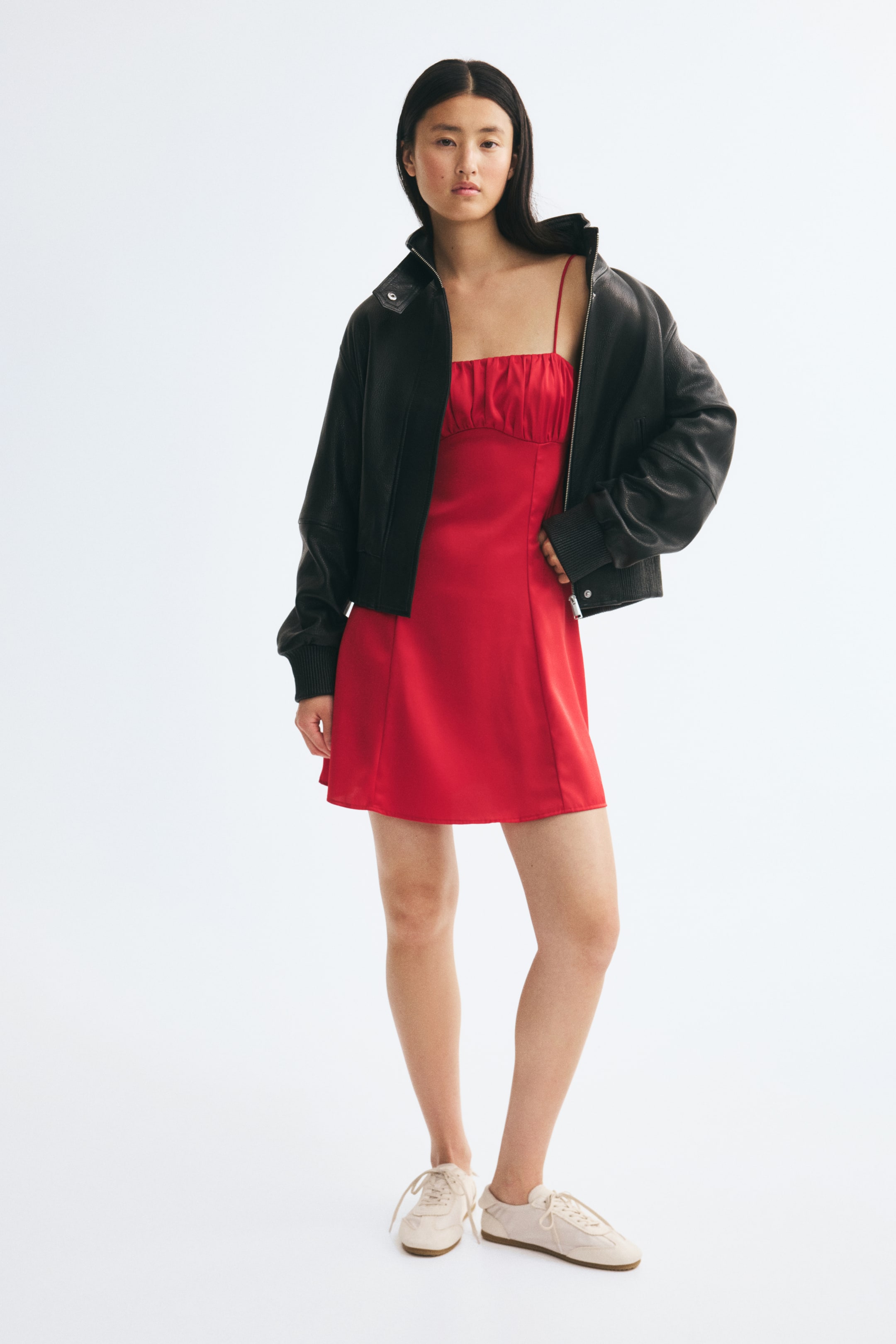 View larger image: A model is wearing a short, bright red dress in a front-facing view, featuring a straight neckline with a gathered, ruched bust and thin spaghetti straps. The dress has visible vertical seams running down the front of its gently flared skirt and is styled with an oversized black leather jacket and off-white sneakers.