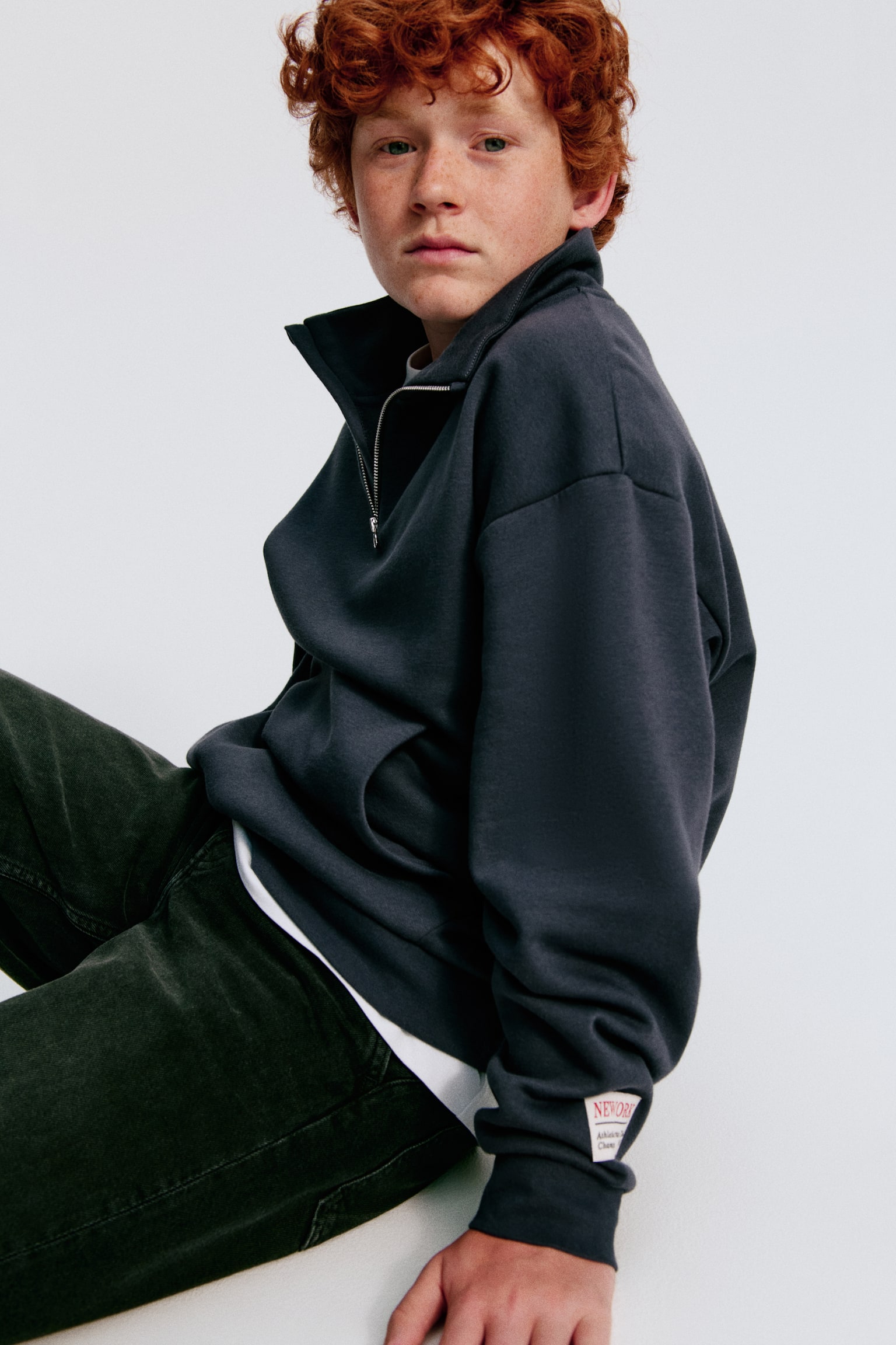Un sweat-shirt gris foncé est présenté à plat, face avant, avec un col montant et une fermeture éclair noire courte s'étendant depuis le haut. Il est conçu avec des épaules tombantes, des manches longues, une grande poche kangourou à l'avant, et des finitions en bord-côte aux poignets et à l'ourlet. - 1