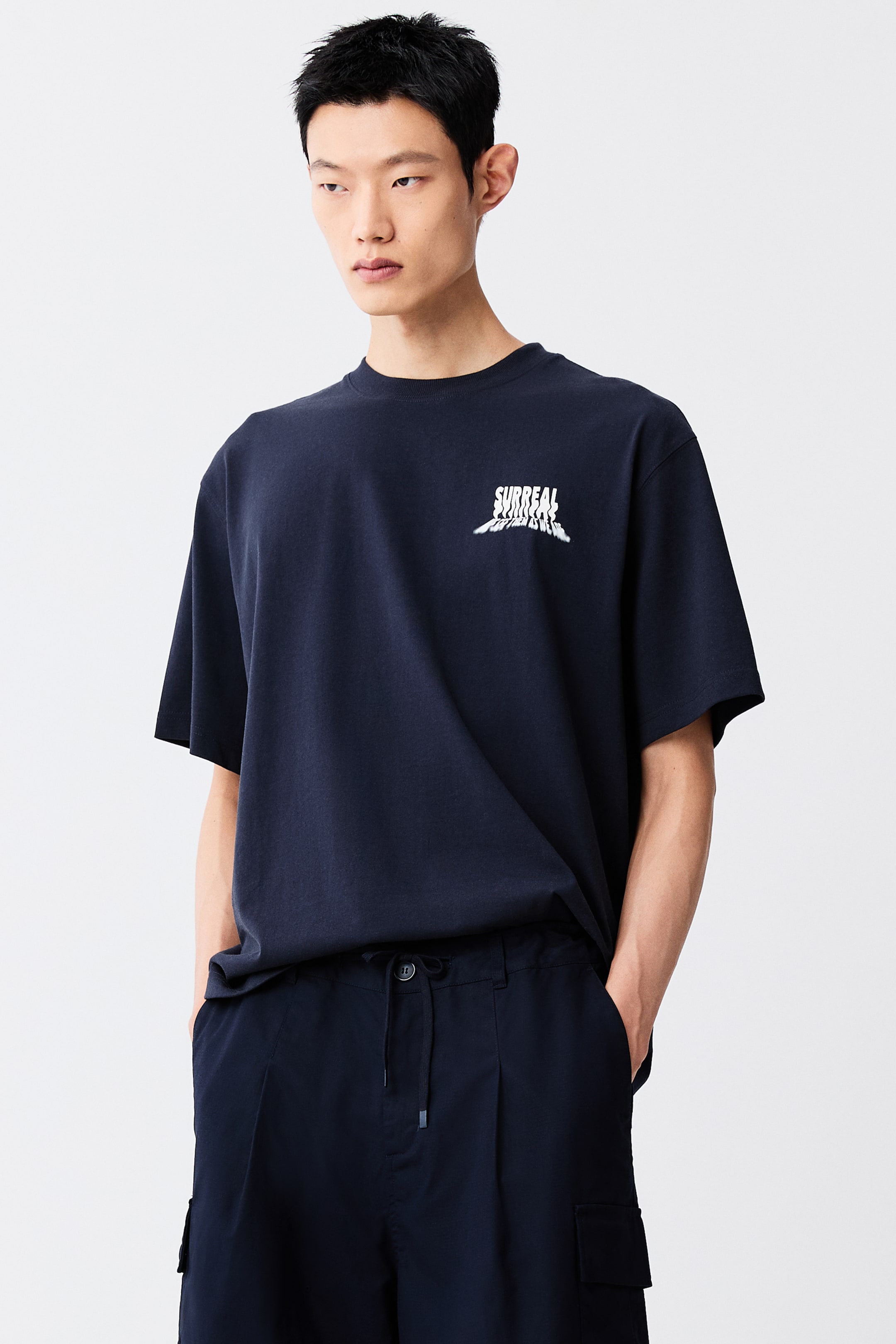 View larger image: A dark blue heavyweight cotton jersey T-shirt is worn by the model, featuring a white 'SURREAL FUTURE' graphic print on the chest. It is designed with a round rib-trimmed neckline, dropped shoulders, and a straight-cut hem, giving it a loose silhouette.